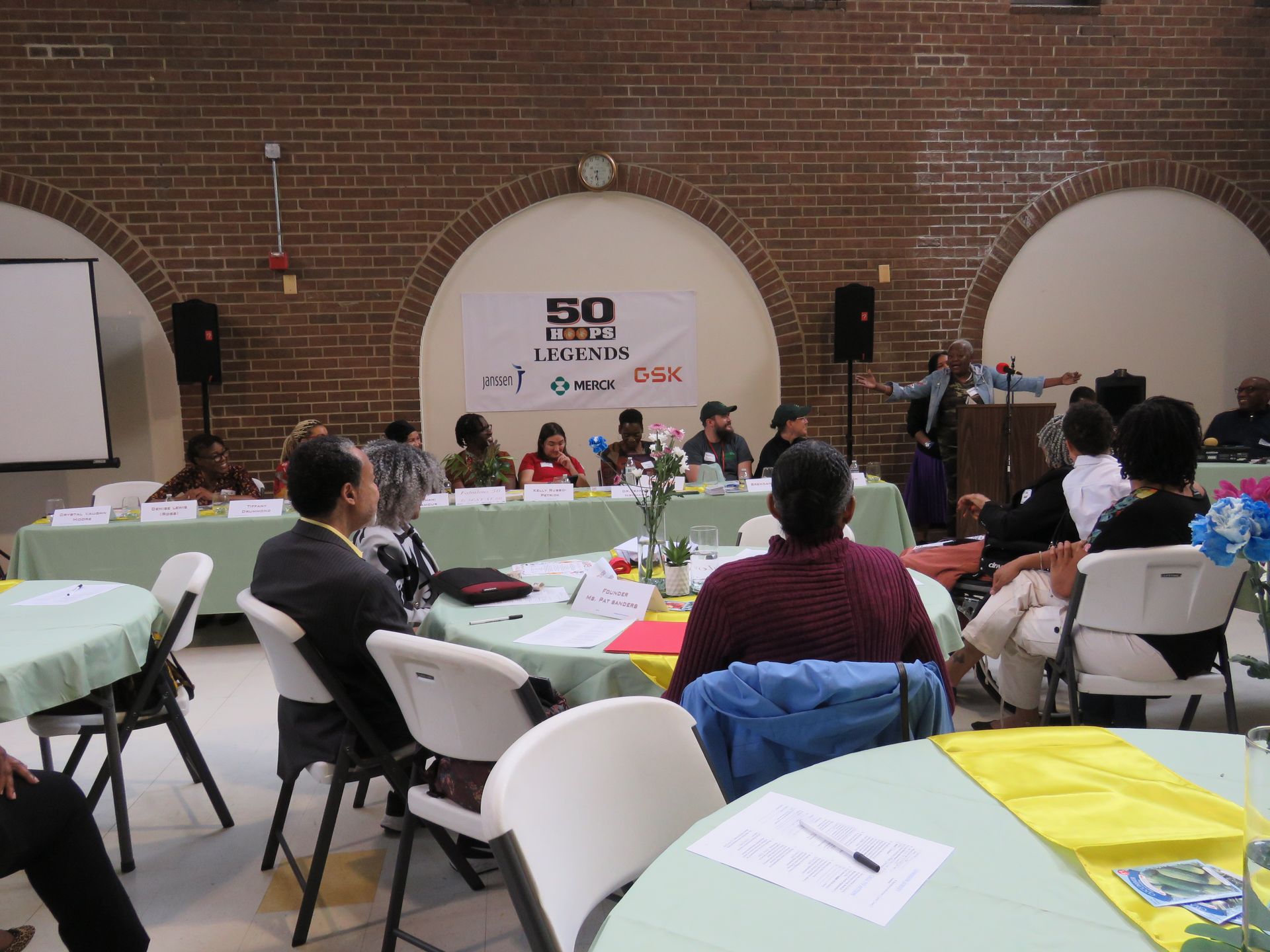 Panel discussion at a conference, with attendees at round tables and panelists at a long table under arches.