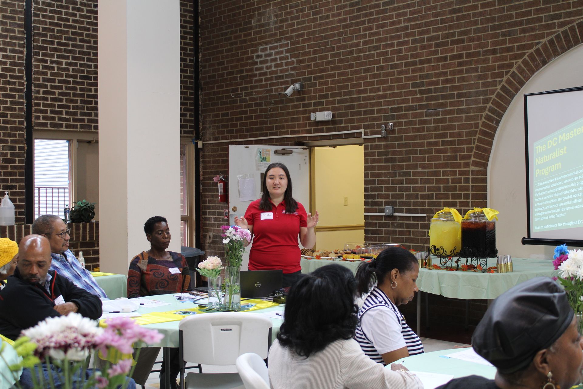 Woman in red top presents to group at a table in front of a brick wall and projector screen.