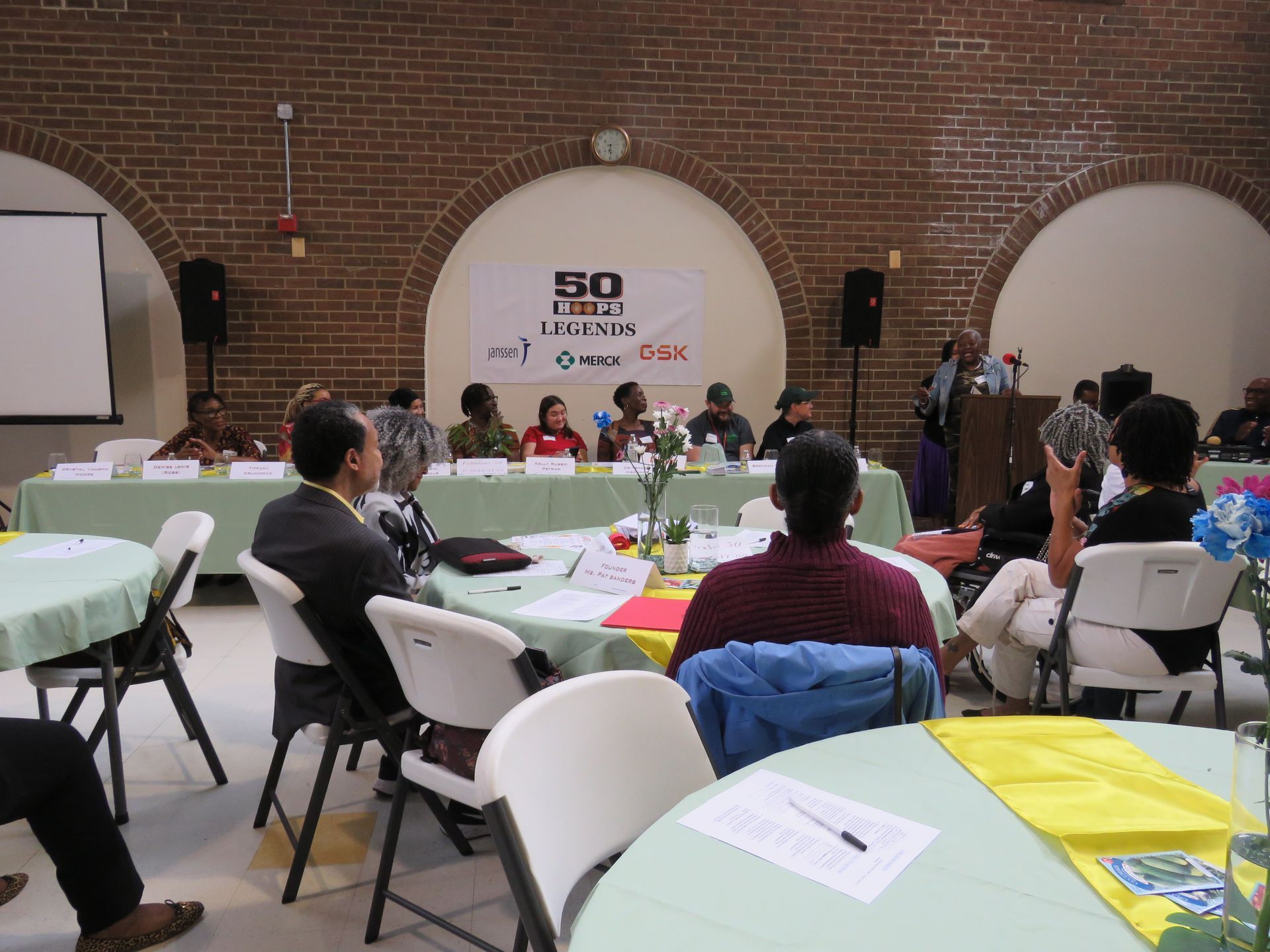 Panel discussion at a conference, speakers seated at a table, audience at round tables in a brick-walled room.