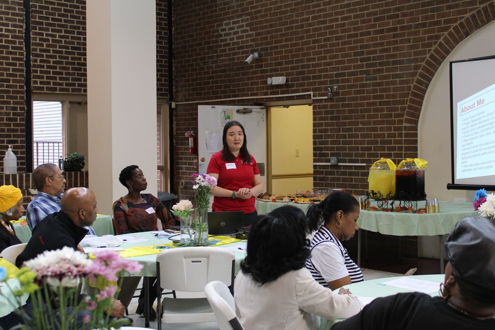 A woman in red shirt presents to a seated group at a table in a brick-walled room.