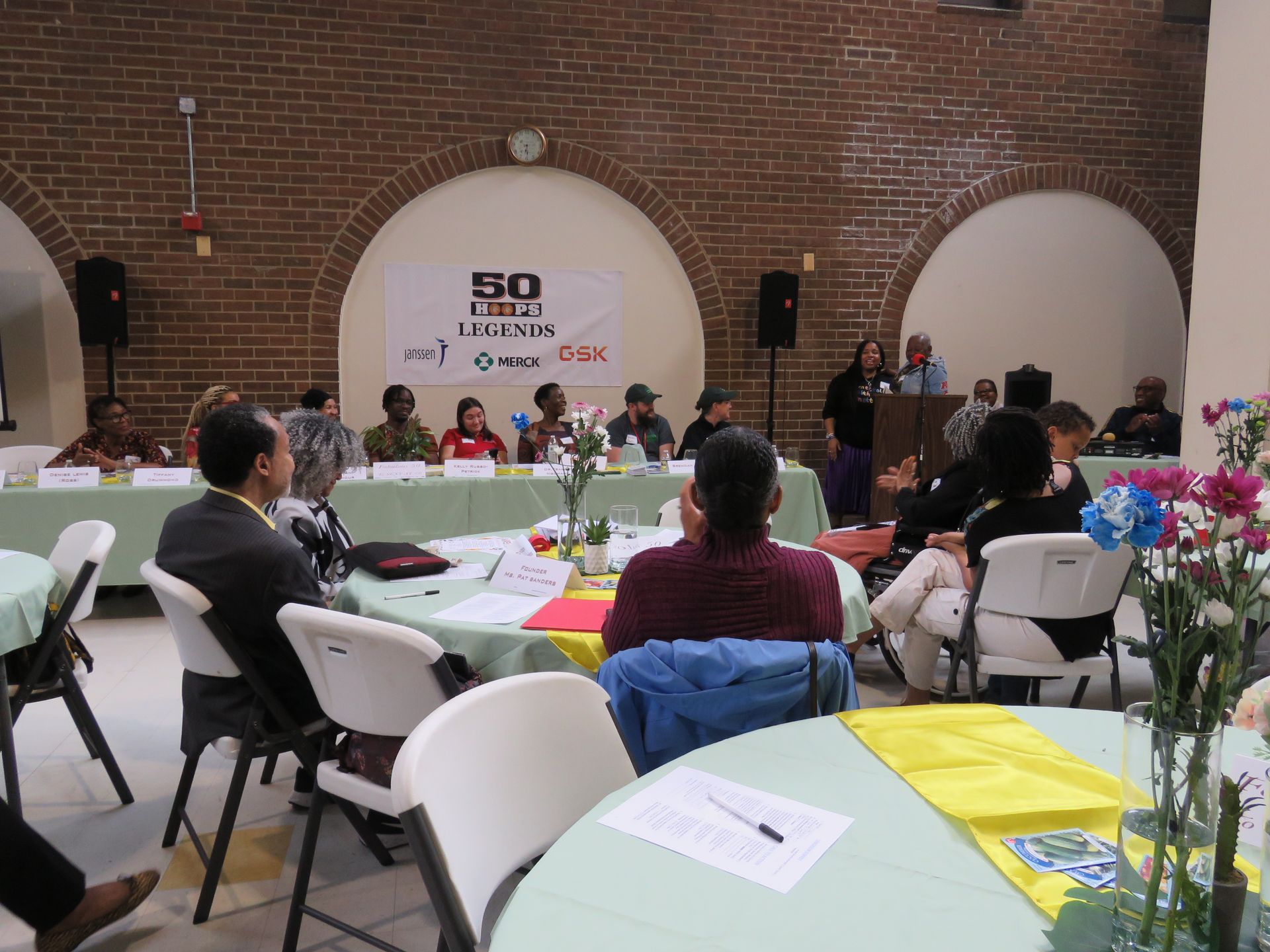 People seated at tables in a room, listening to a presentation on a stage; a banner says “50 Years Undeniable.”