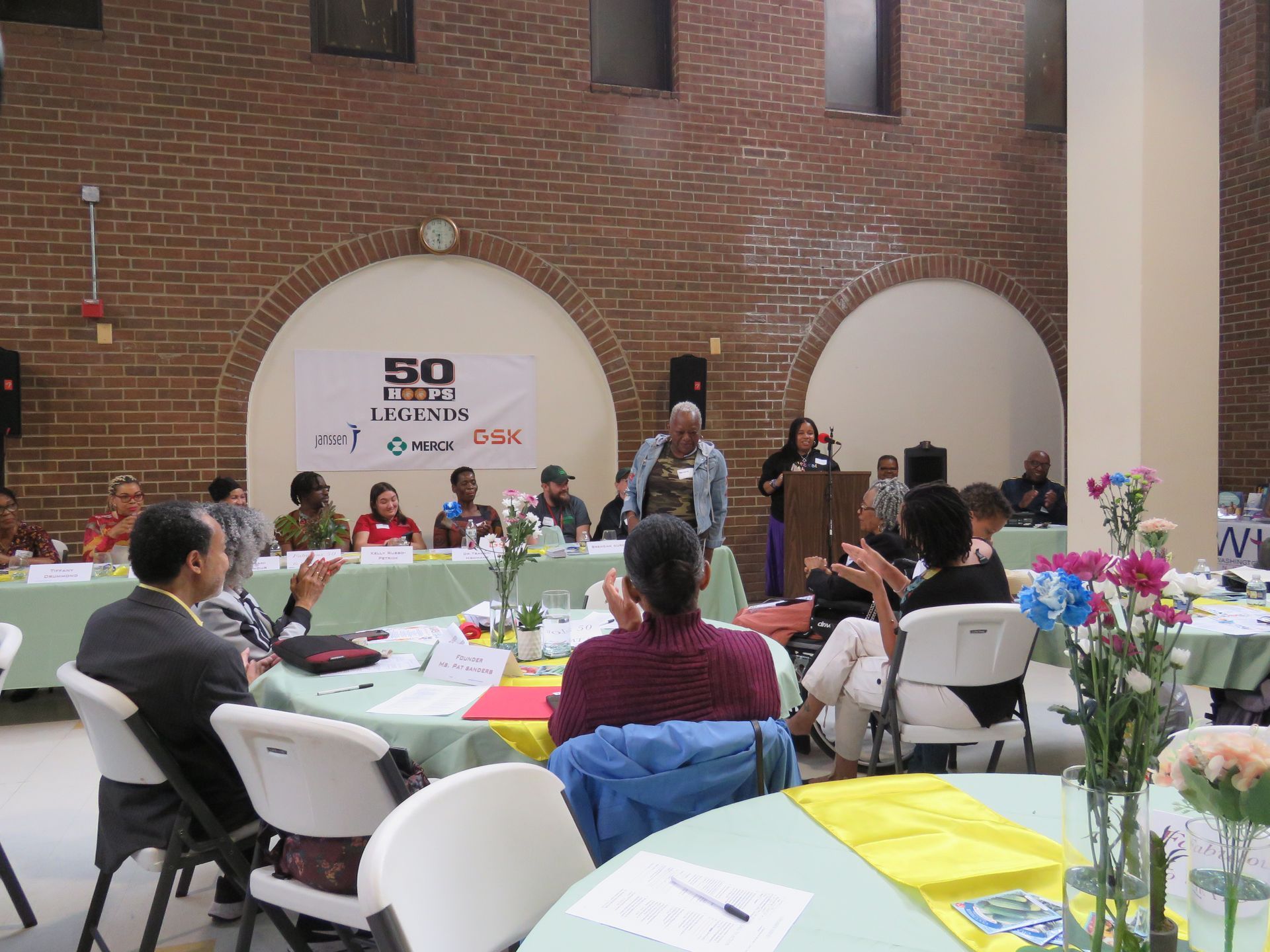 People at a conference with a speaker at a podium, clapping audience, and tables with green tablecloths.