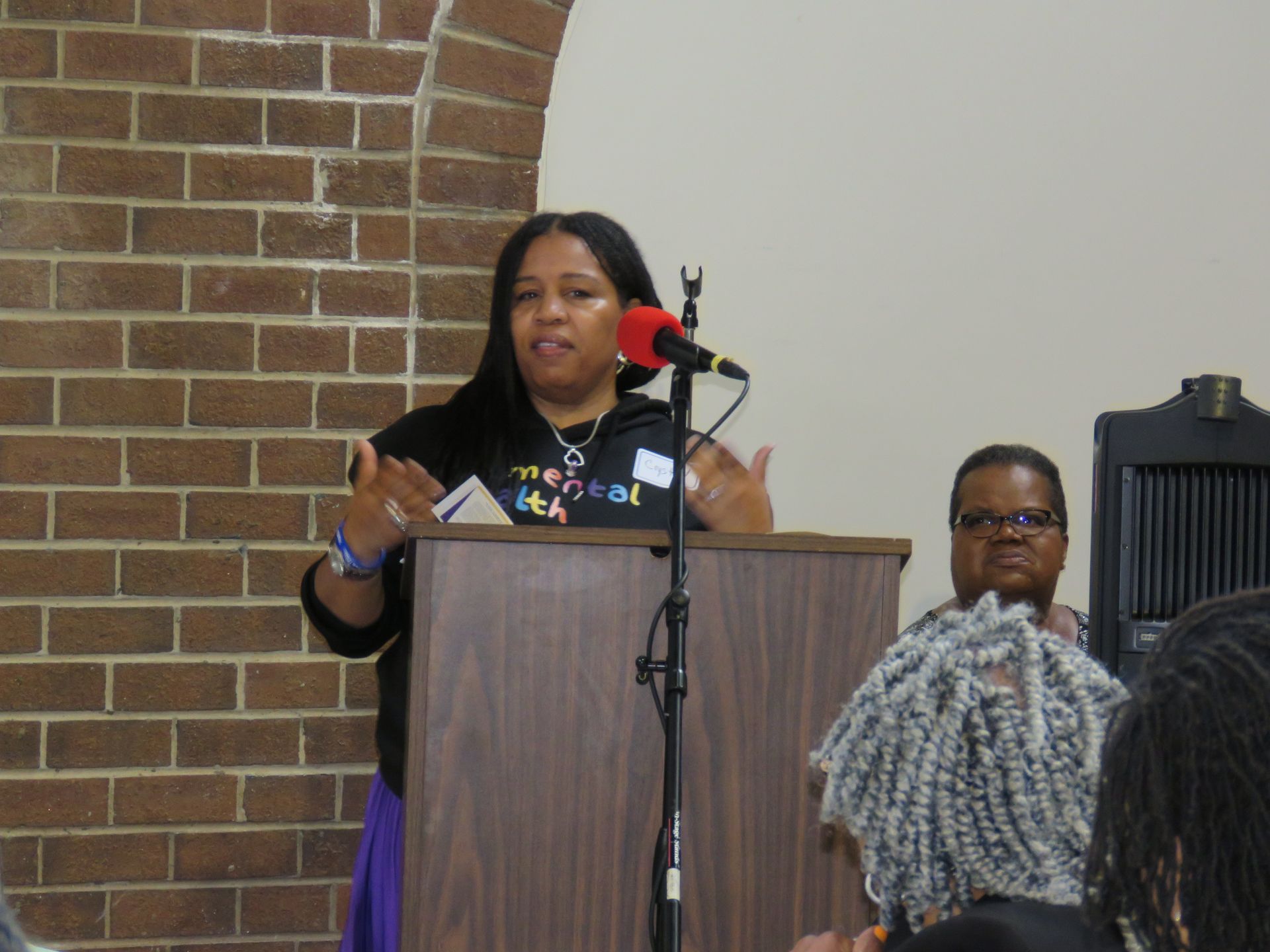 Woman speaking at a podium, gesturing with her hands. Brick wall on the left, audience seated in front.