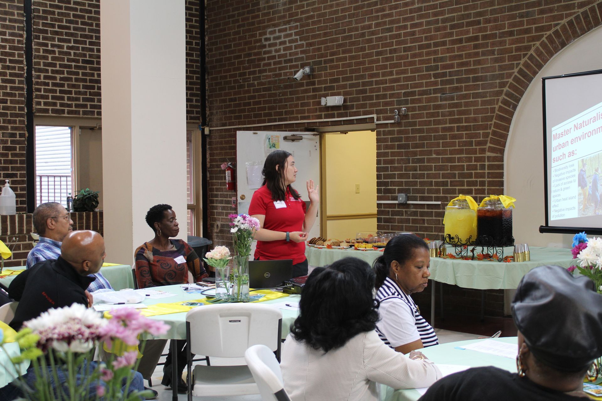 A woman presents to a seated audience at a meeting. Brick wall, projector screen, and refreshments.