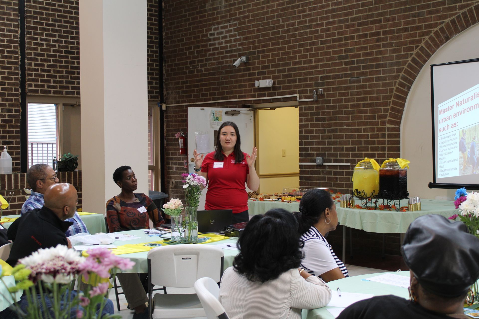 Woman presenting in a room with attendees, projector screen, tables with flowers, and snacks.