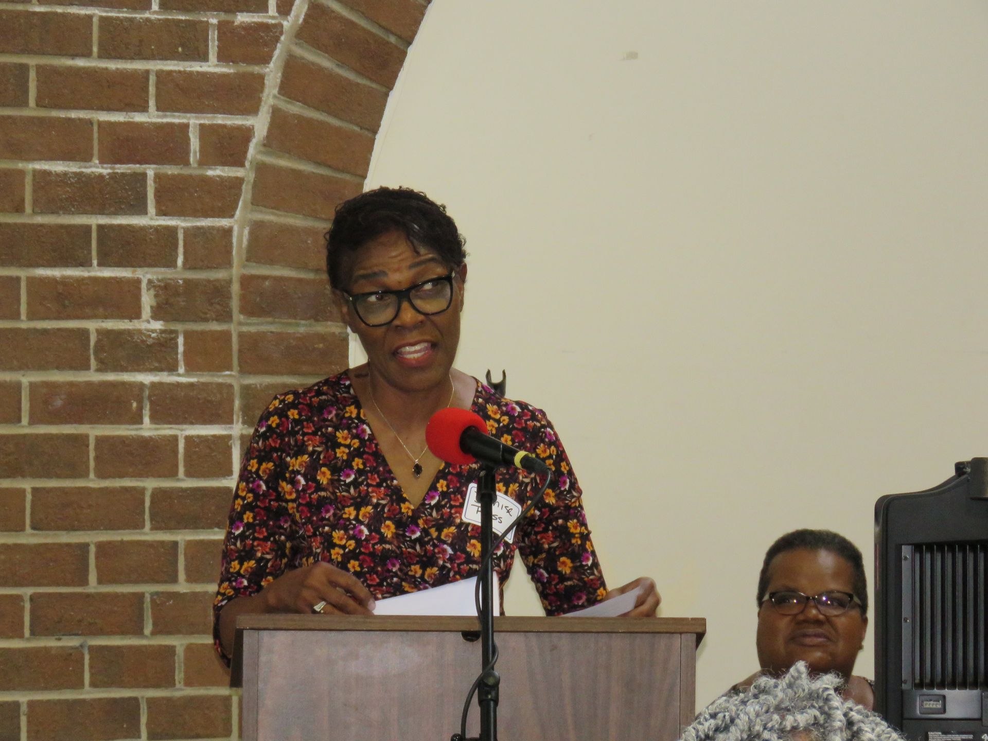 Woman speaking at a podium with microphone in front of brick wall. Another person is seated.