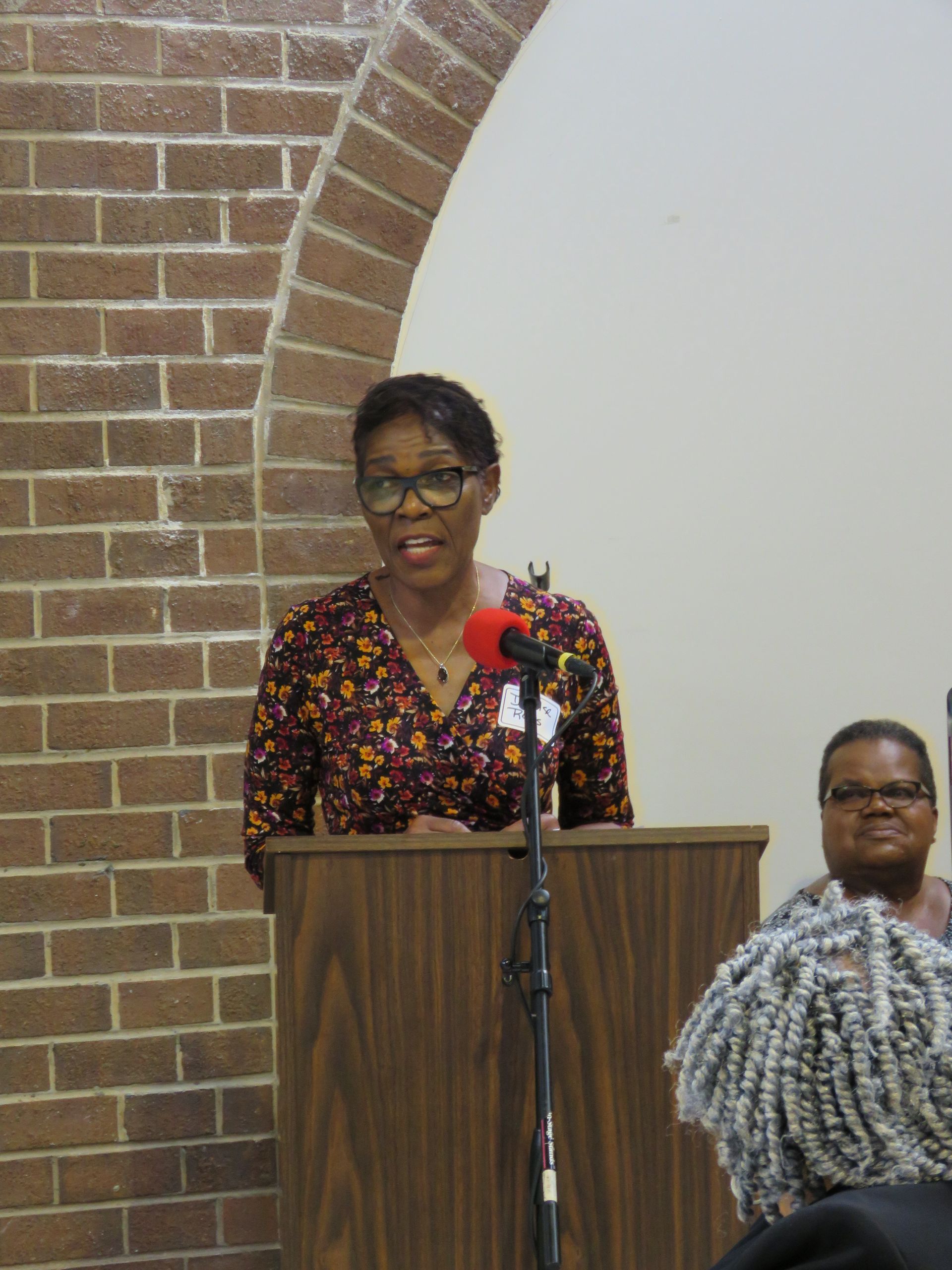 Woman speaking at a podium with microphone, in front of brick arch and audience member.