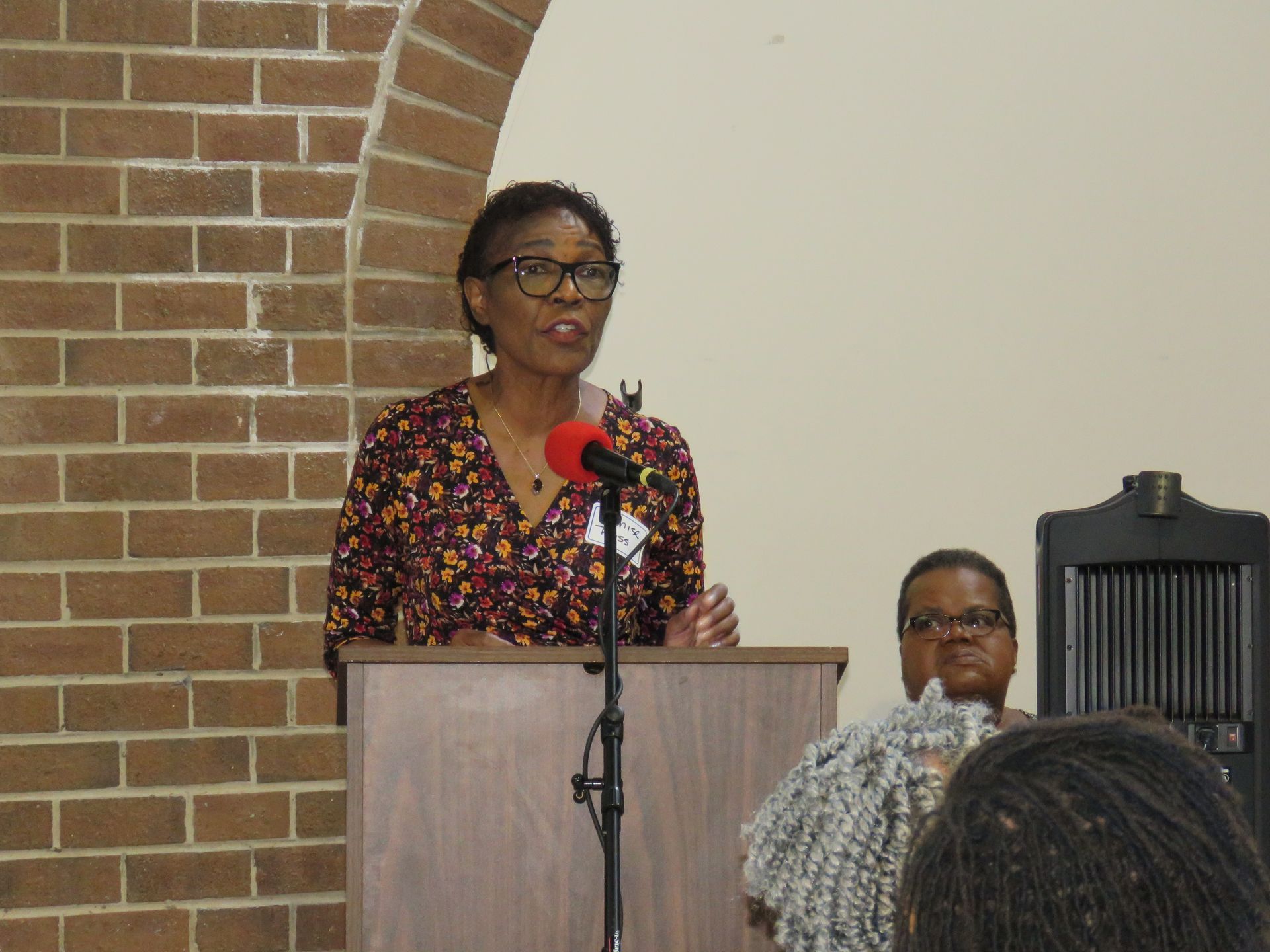 Woman speaking at a lectern, microphone present. Brick wall backdrop. Another person partially visible to the right.