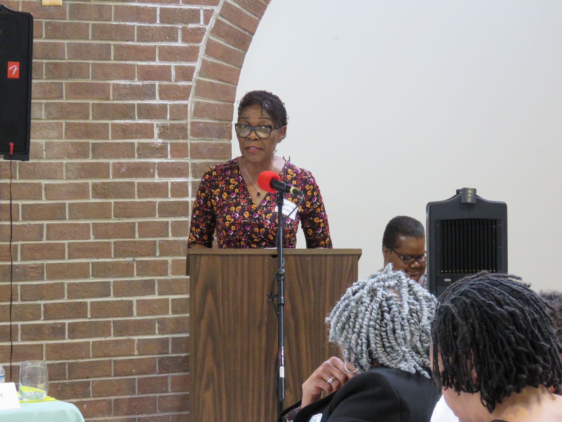 Woman speaking at a podium in a room with a brick wall. Audience members are seated.