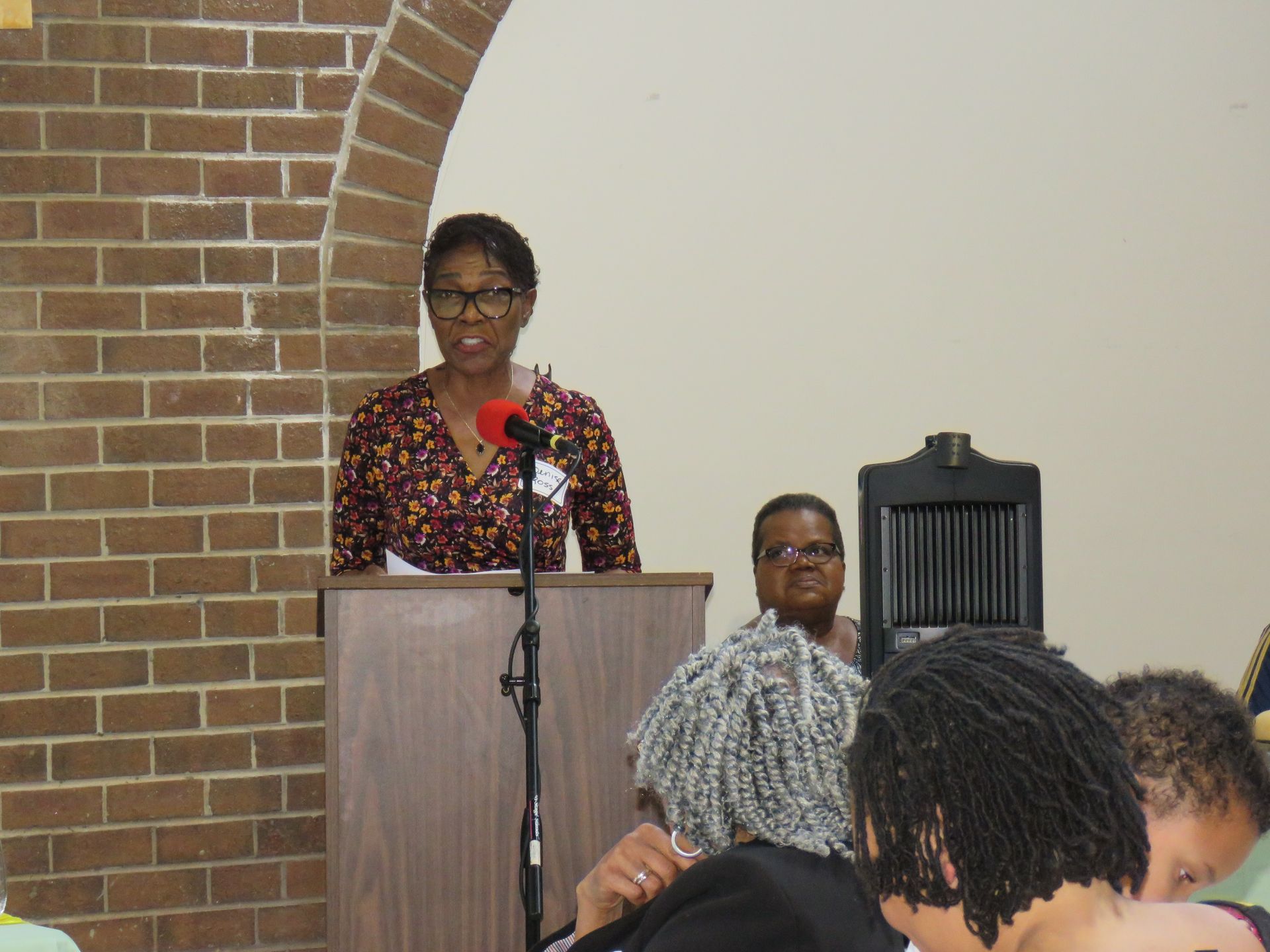 Woman speaking at a podium in a room with an audience. Brick archway and a wooden pulpit.