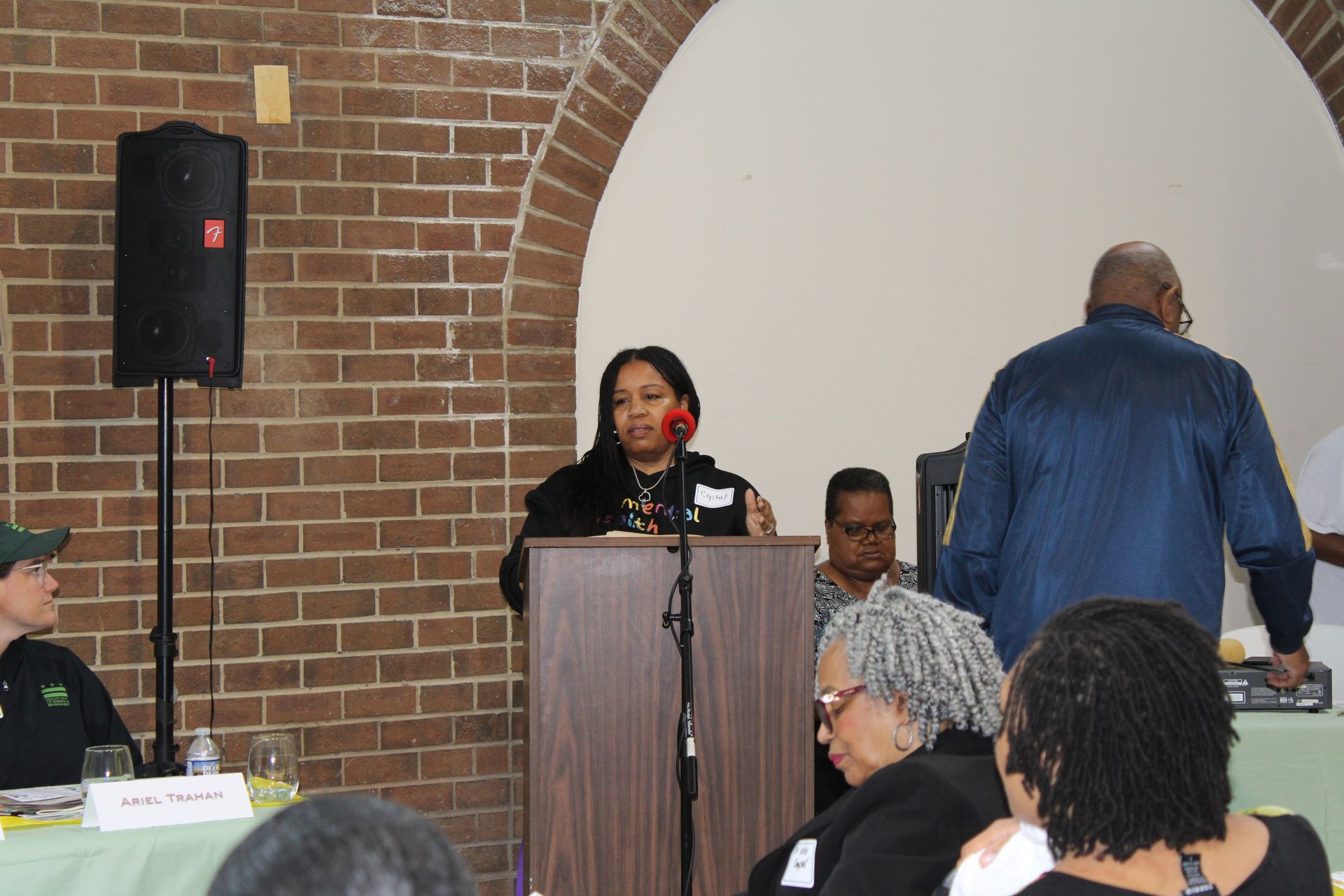 Woman speaking at a podium; brick wall background, people seated at tables, speaker on the left.
