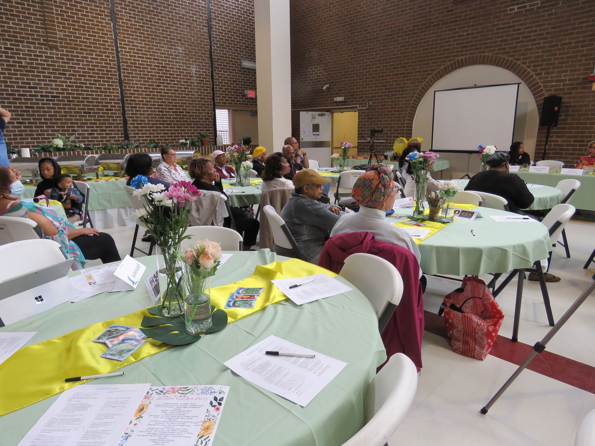People seated at round tables in a decorated room, attending an event with a projector screen.