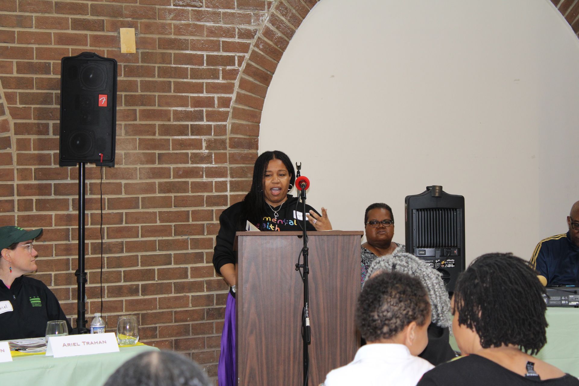 Woman speaking at a podium, in a brick-walled room with a panel of seated people.