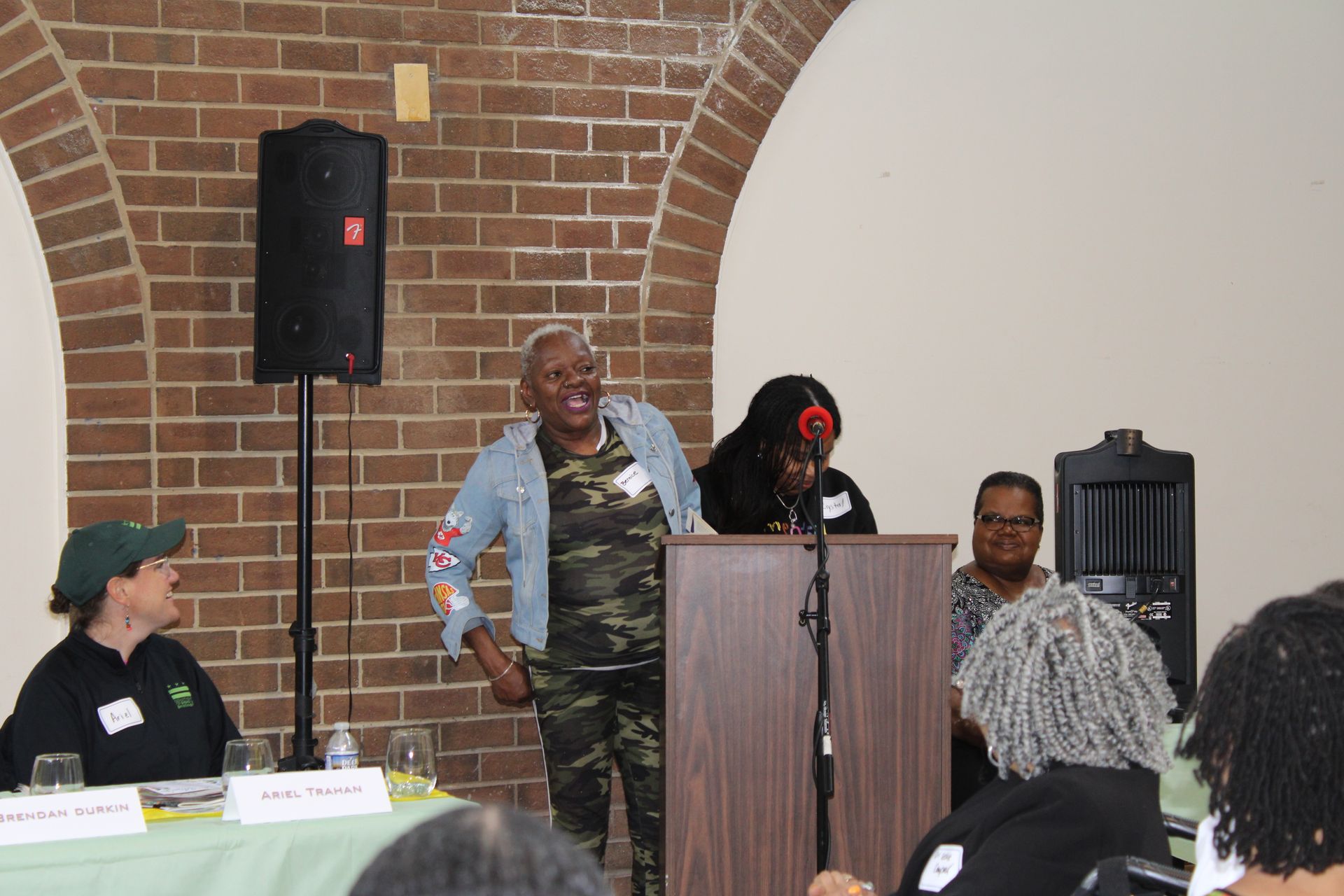 Panel discussion at a brick-walled venue. Woman in camo speaks. Others listen, some at a table with nameplates.