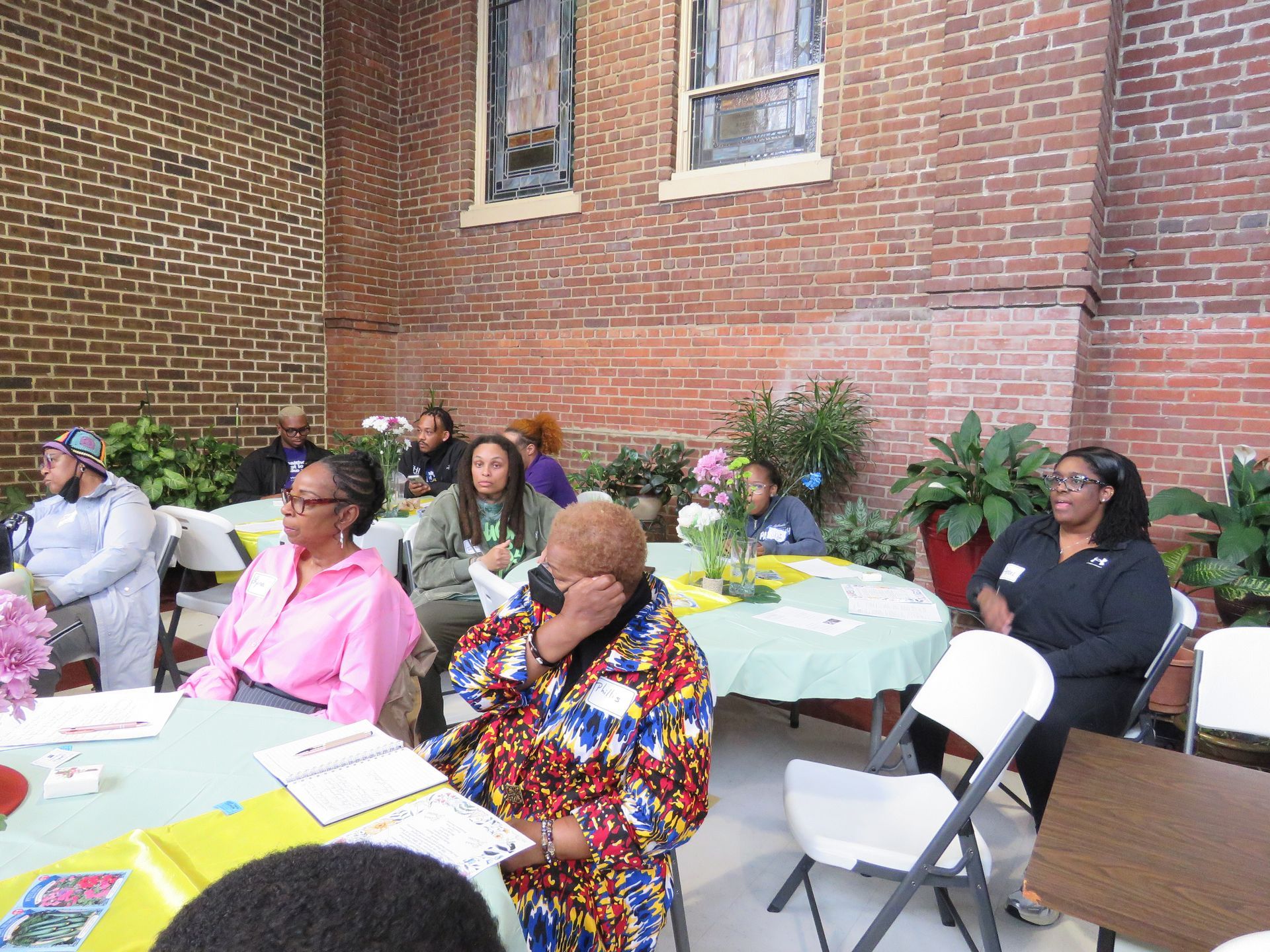 People seated at round tables in an outdoor space with brick walls and flowers, listening.