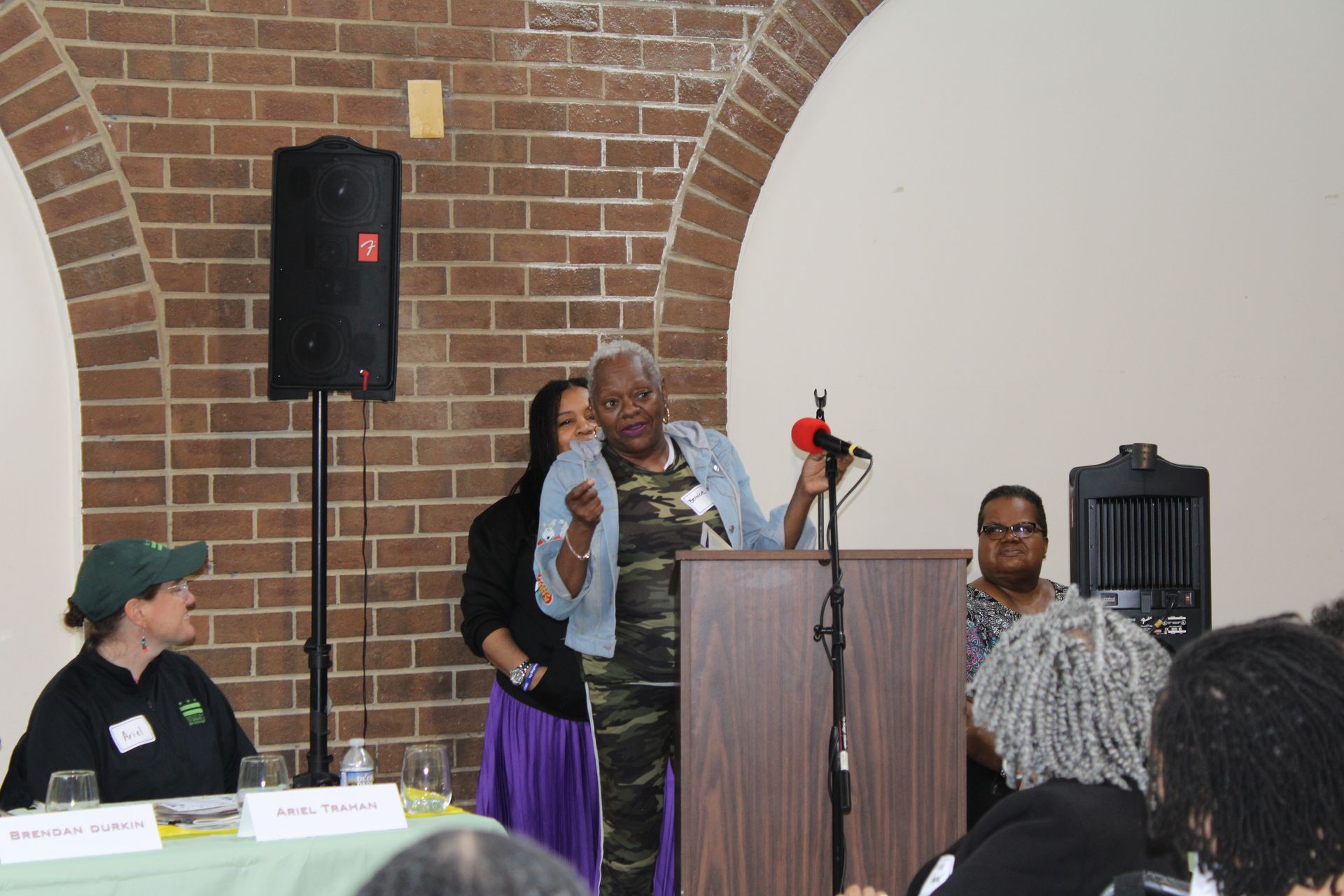 Woman speaking at a podium, others seated behind a table in front of a brick wall and arches.