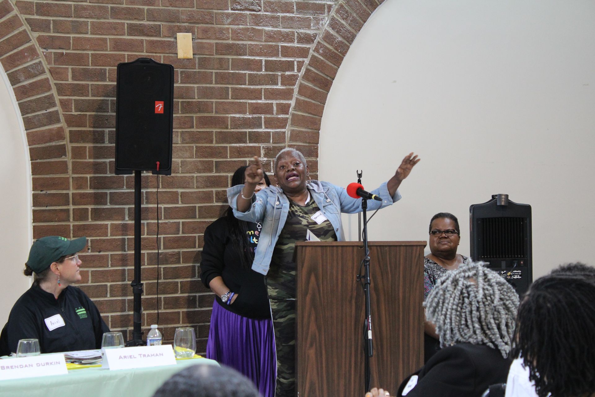Woman speaking passionately at a podium, arms raised, in a room with a brick wall backdrop.