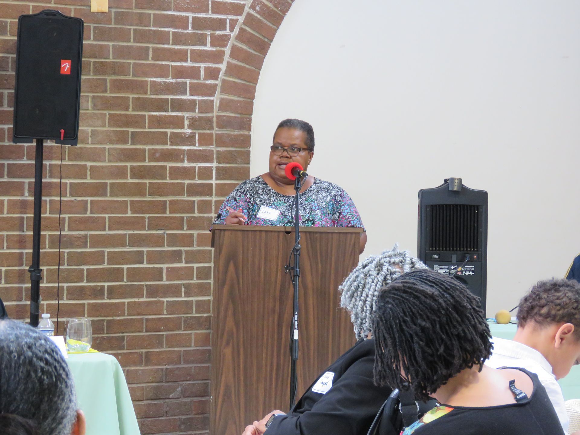 Woman speaking at a lectern, facing an audience. Brick wall background, speaker on left.