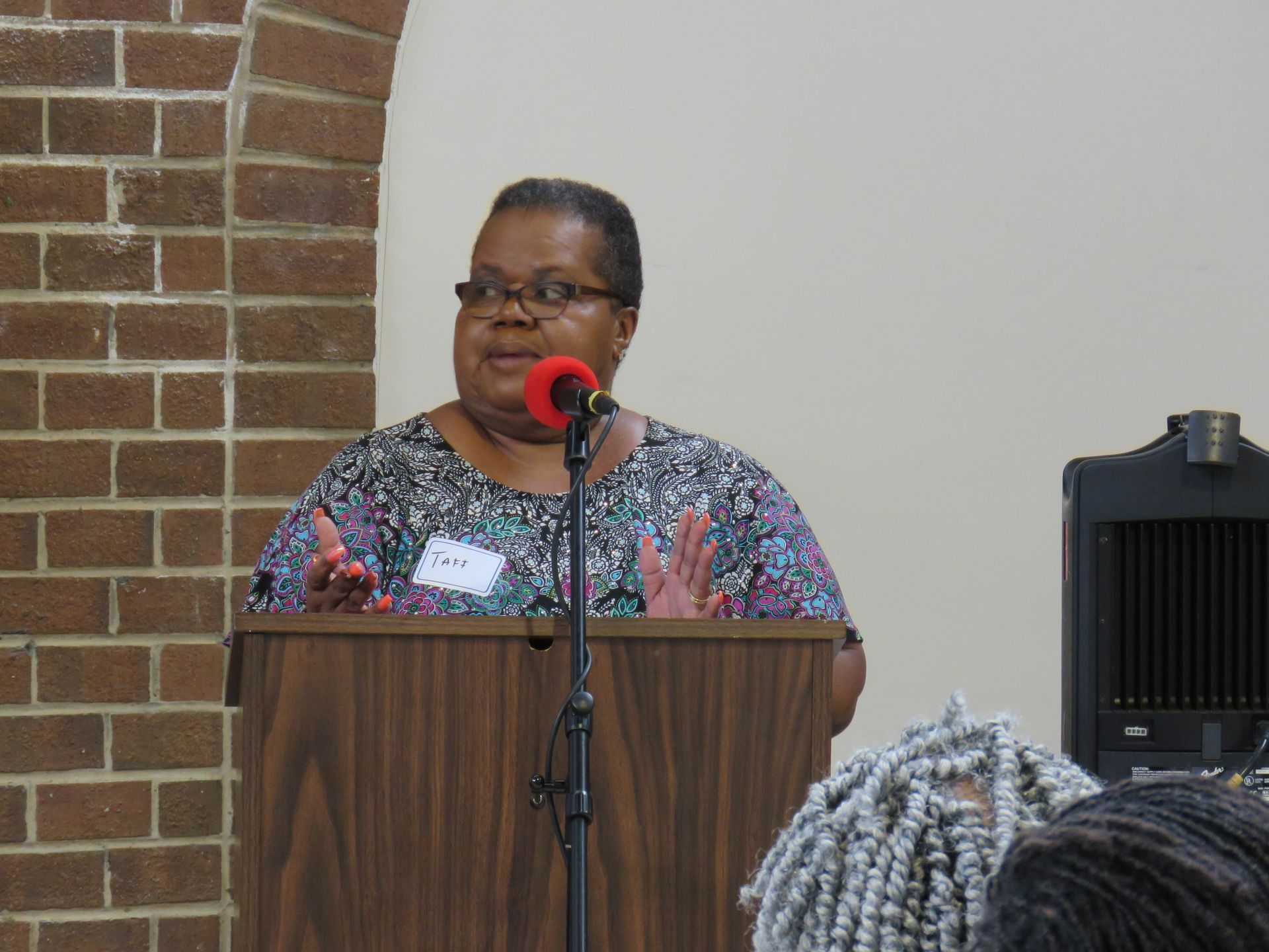 Woman speaking at a lectern, gesturing with hands. Brick wall and speaker system in the background.
