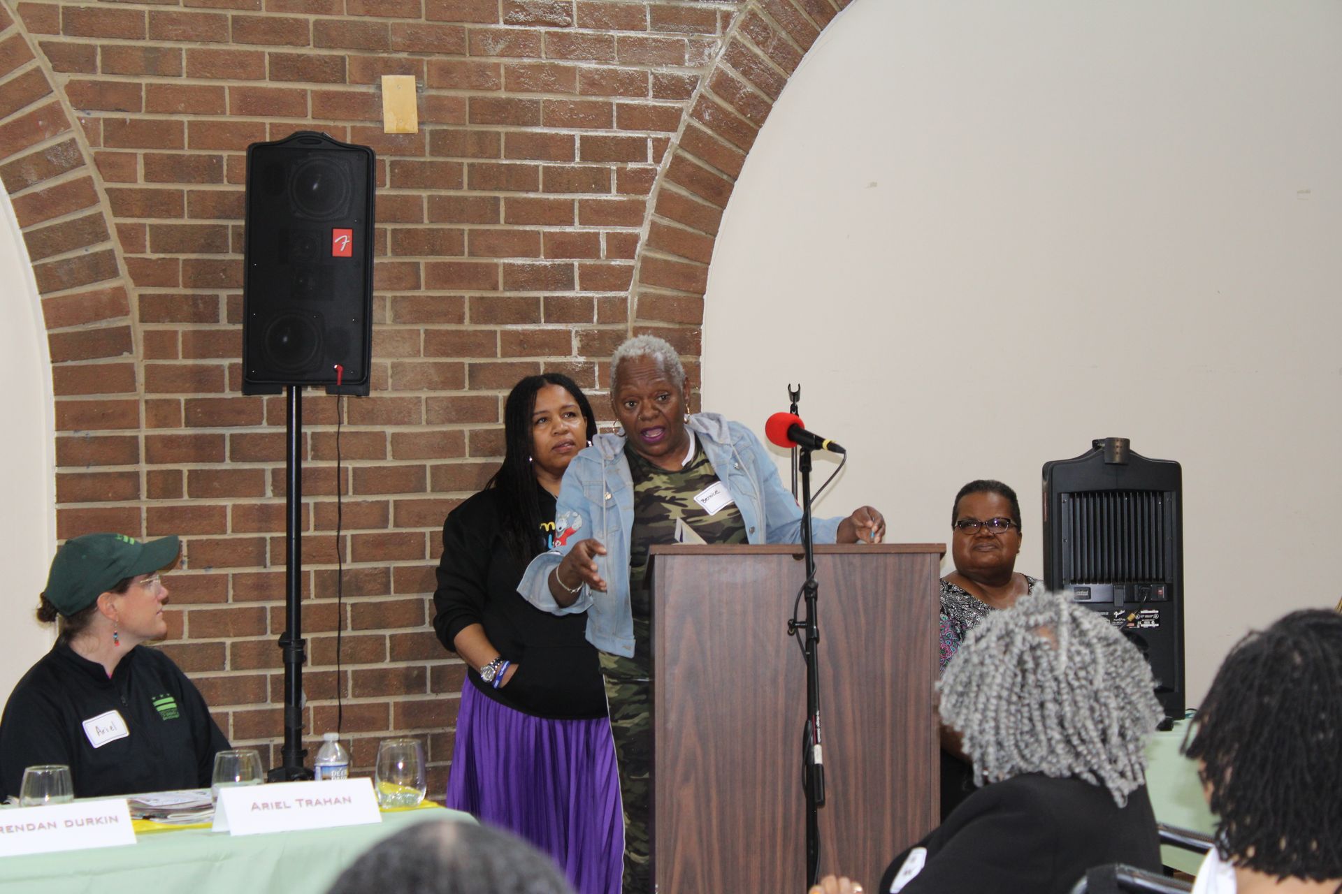 People at a podium, speaking at a meeting. A person in a camouflage jacket gestures. Others sit or stand nearby.