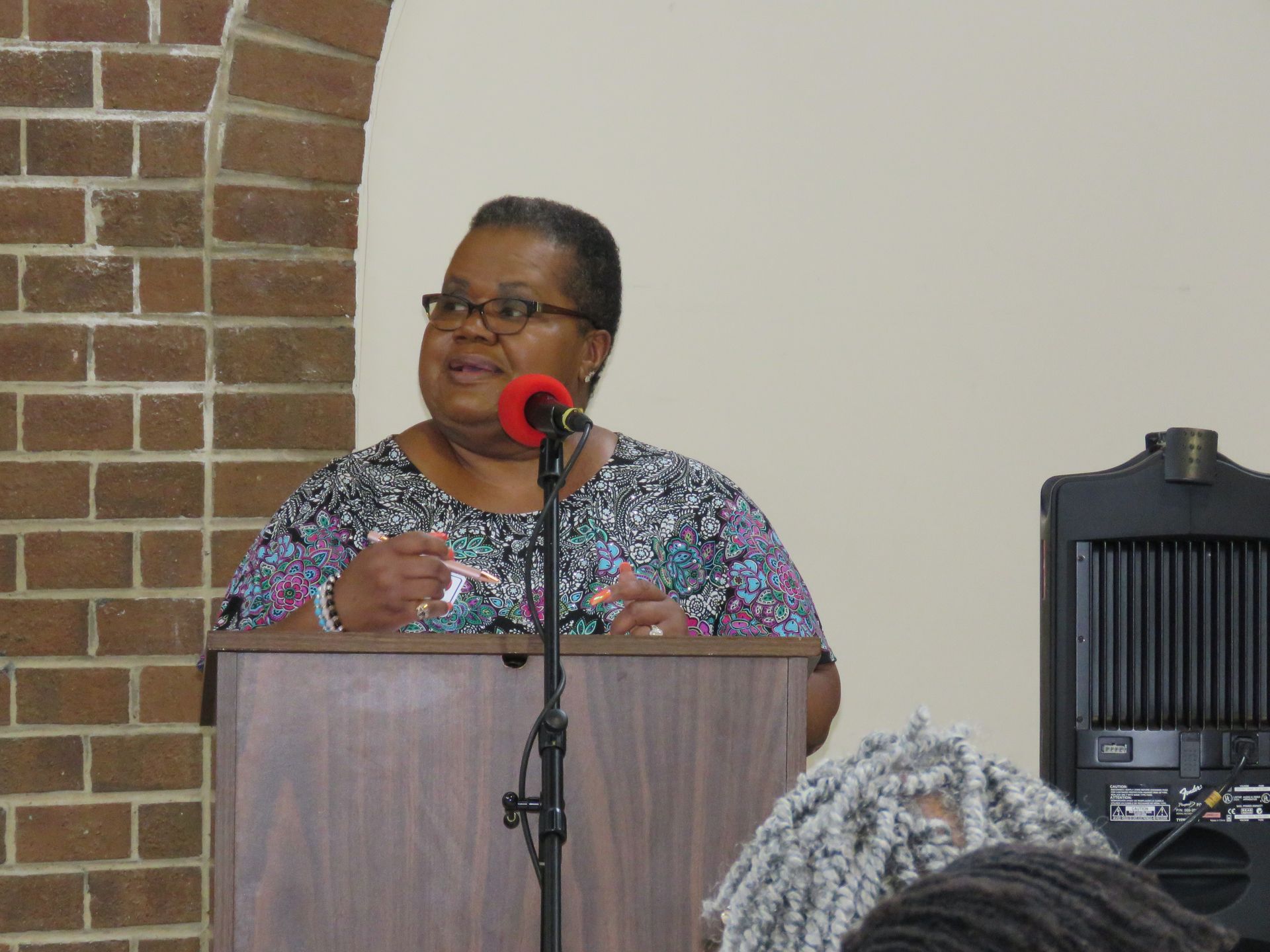 Woman speaking at a podium, wearing glasses, and a patterned shirt, in front of a brick wall.