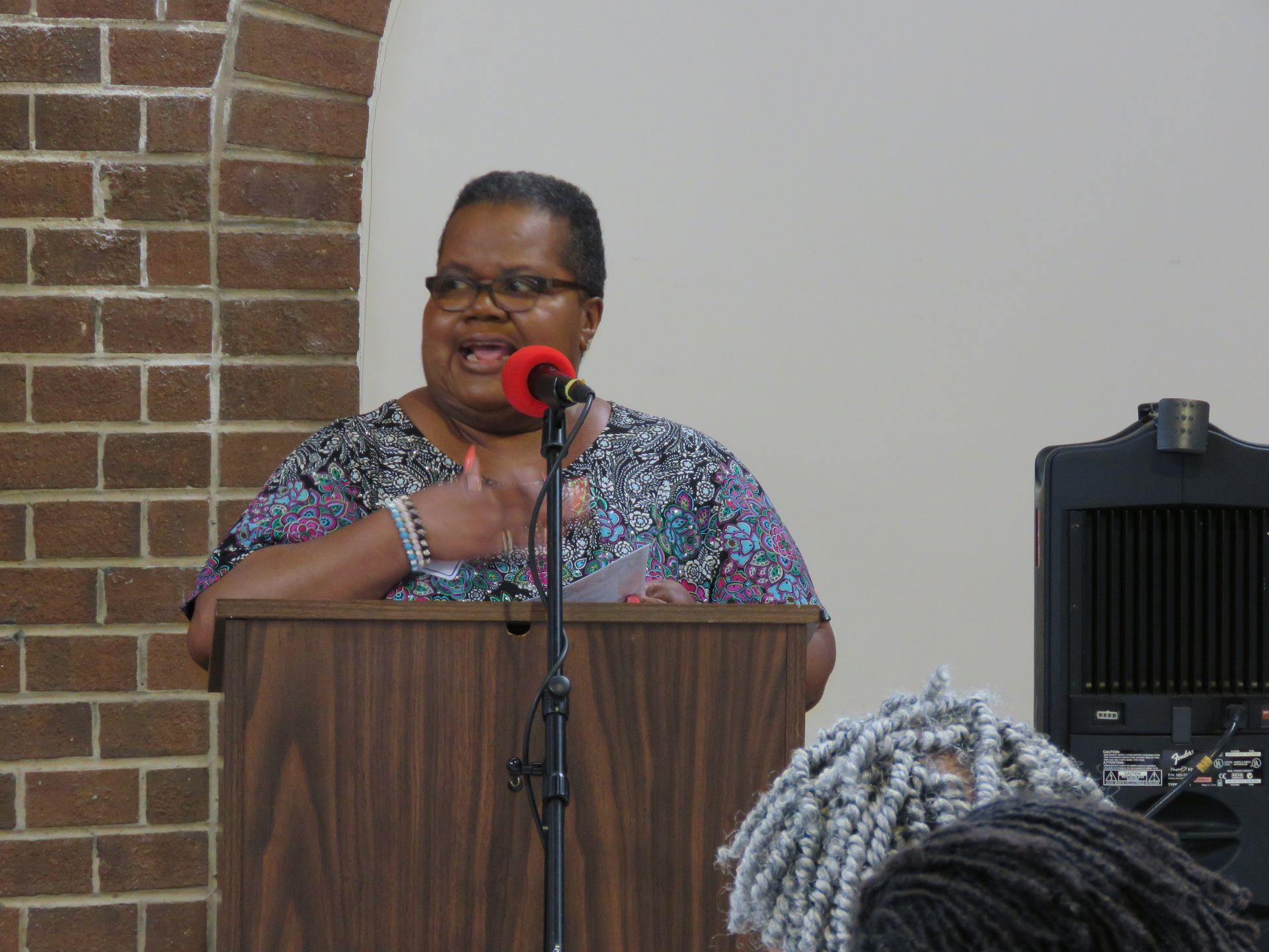 Woman speaking at a podium, red microphone, brick wall background.