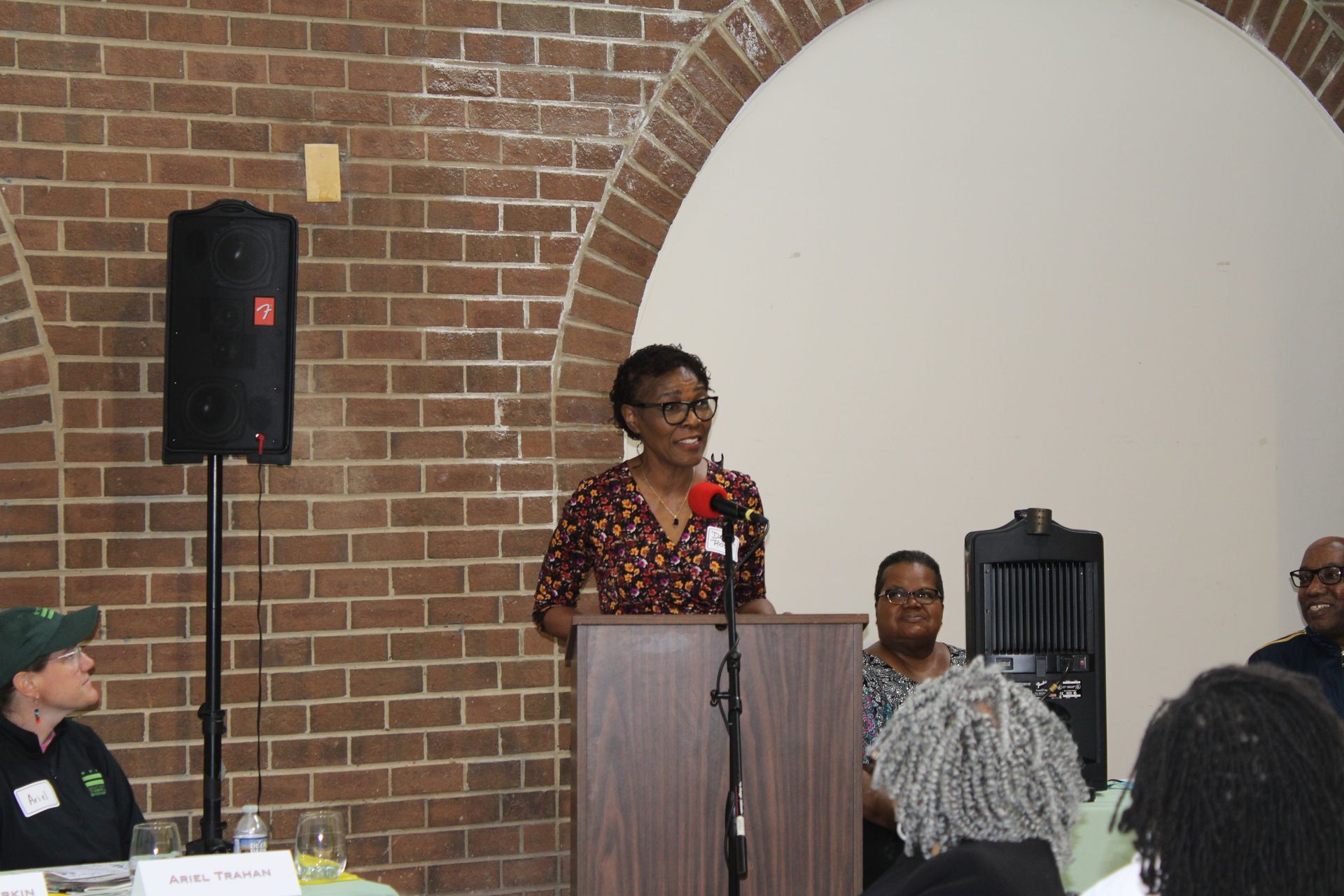 Woman speaking at a podium, brick wall backdrop. Others seated nearby.