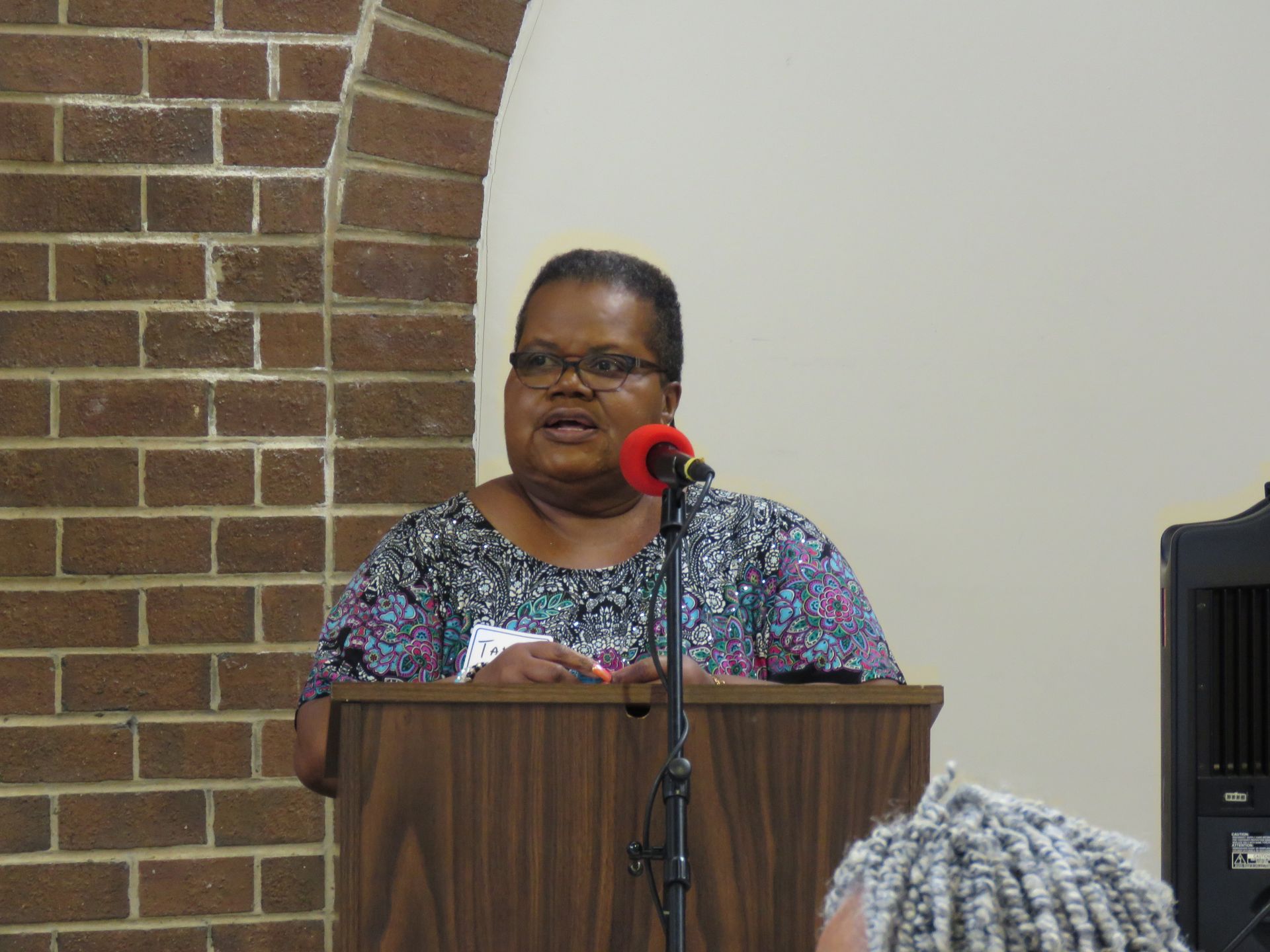 Woman speaking at a podium in front of a brick wall, holding a microphone.