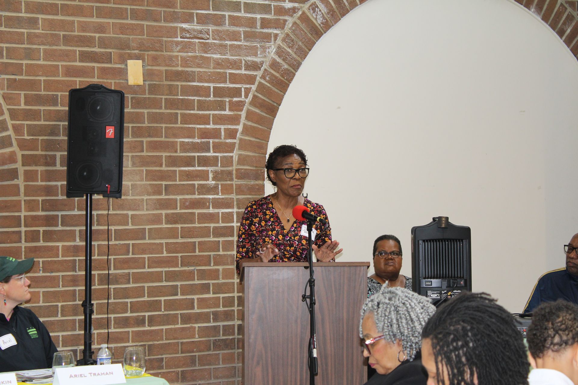 Woman speaking at a podium, brick wall backdrop. People seated, one with grey dreadlocks, listening.