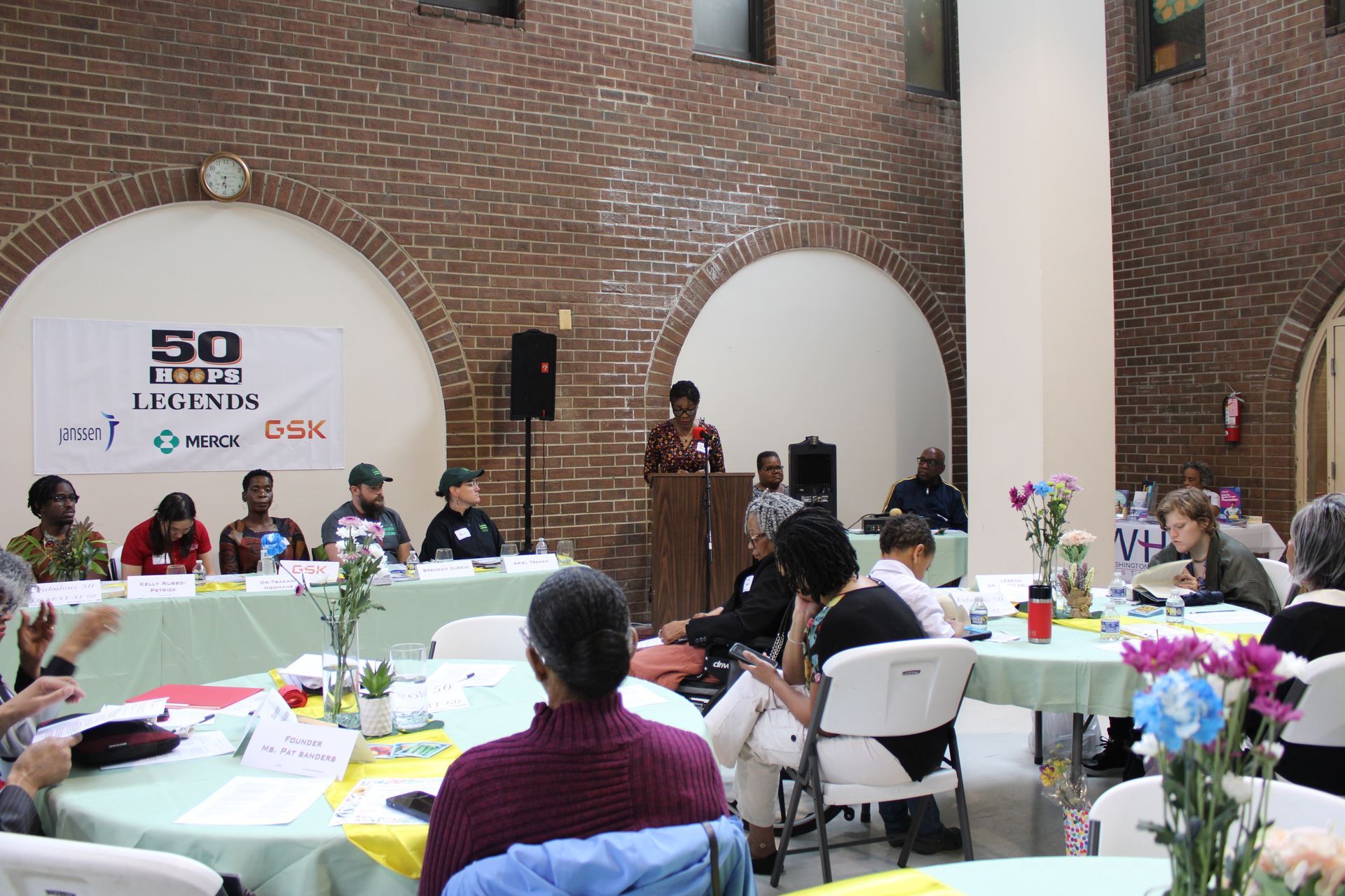 Panel discussion at a conference. A speaker at podium, panel at a table, audience seated at tables. Brick walls.