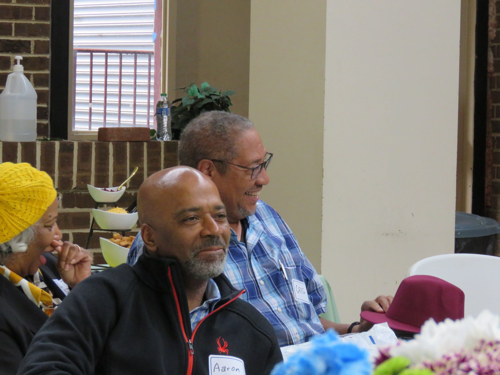 Group of people seated indoors, smiling. A man in black jacket is in the foreground. Brick wall and window visible.
