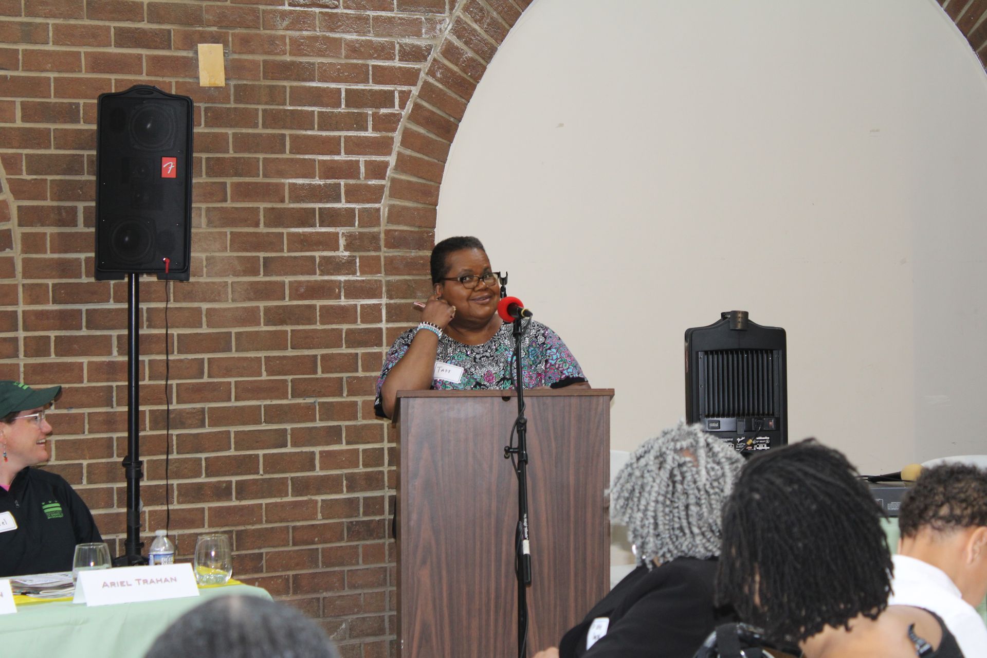 Woman speaking at a podium in a brick-walled room; a microphone is present.