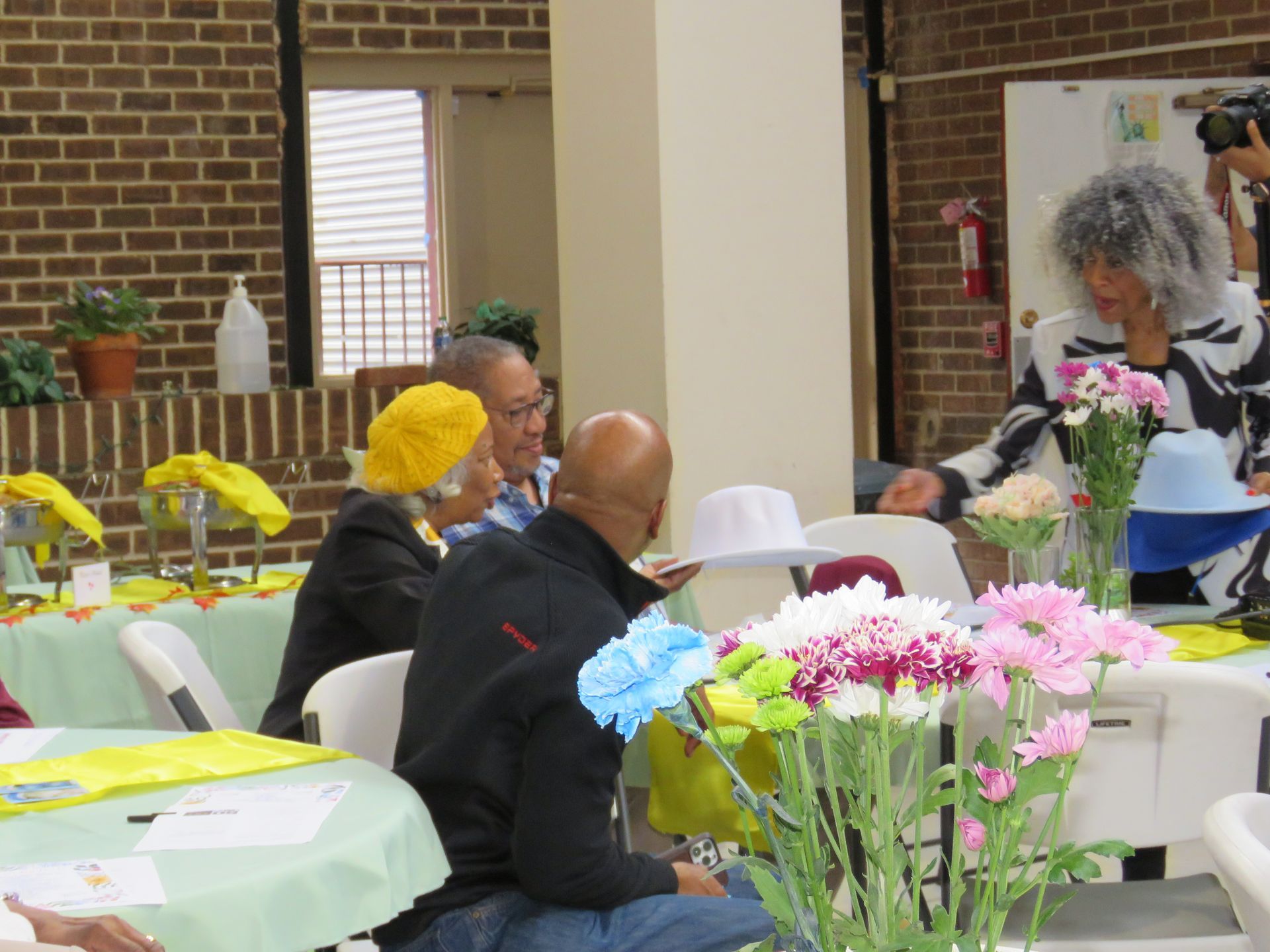 People at a table with flowers, one woman wearing a black and white patterned jacket.