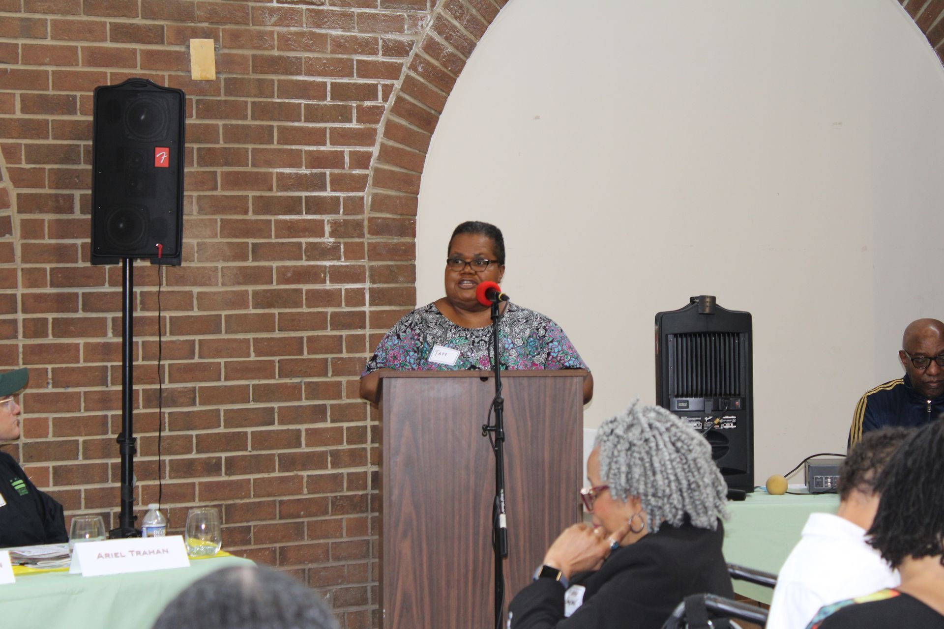 Woman speaking at a podium in a room with a brick wall and arches.