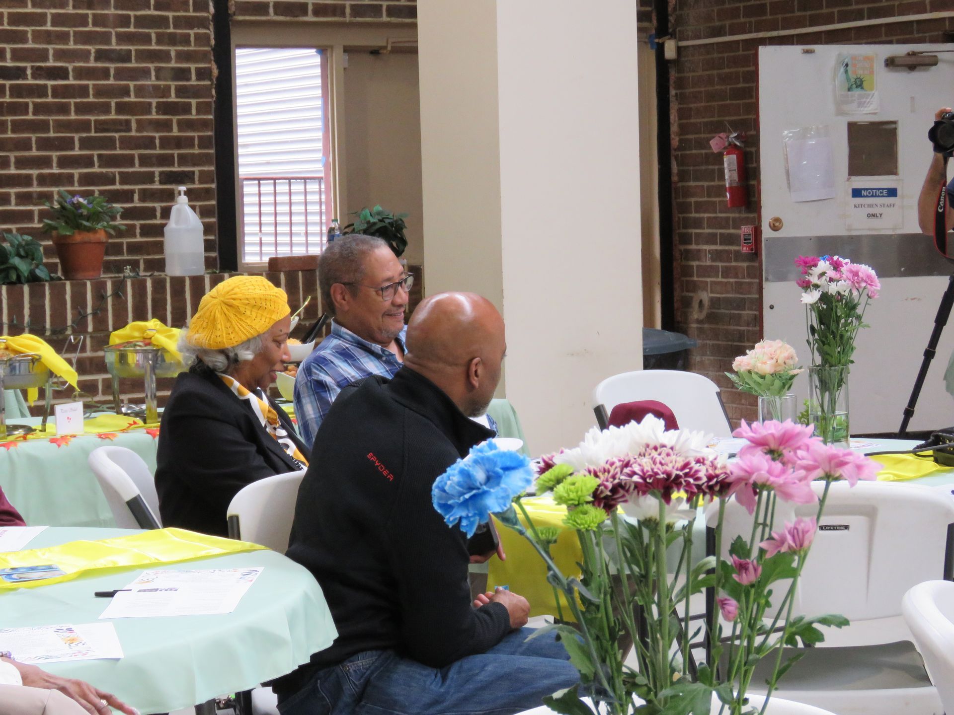 People seated at a table with flowers. Brick wall backdrop.