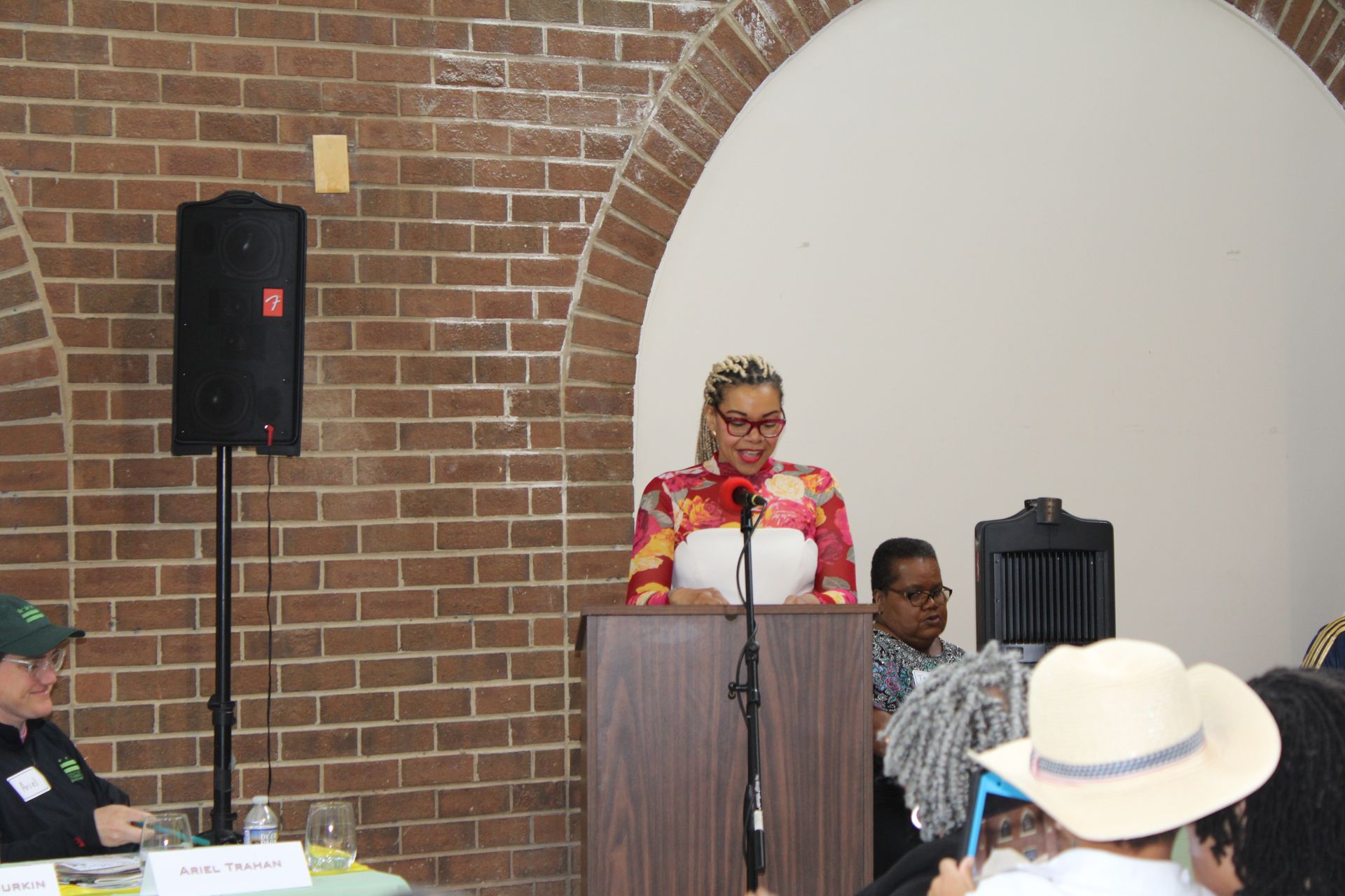 Woman speaking at a podium in a room with brick wall, other people seated.