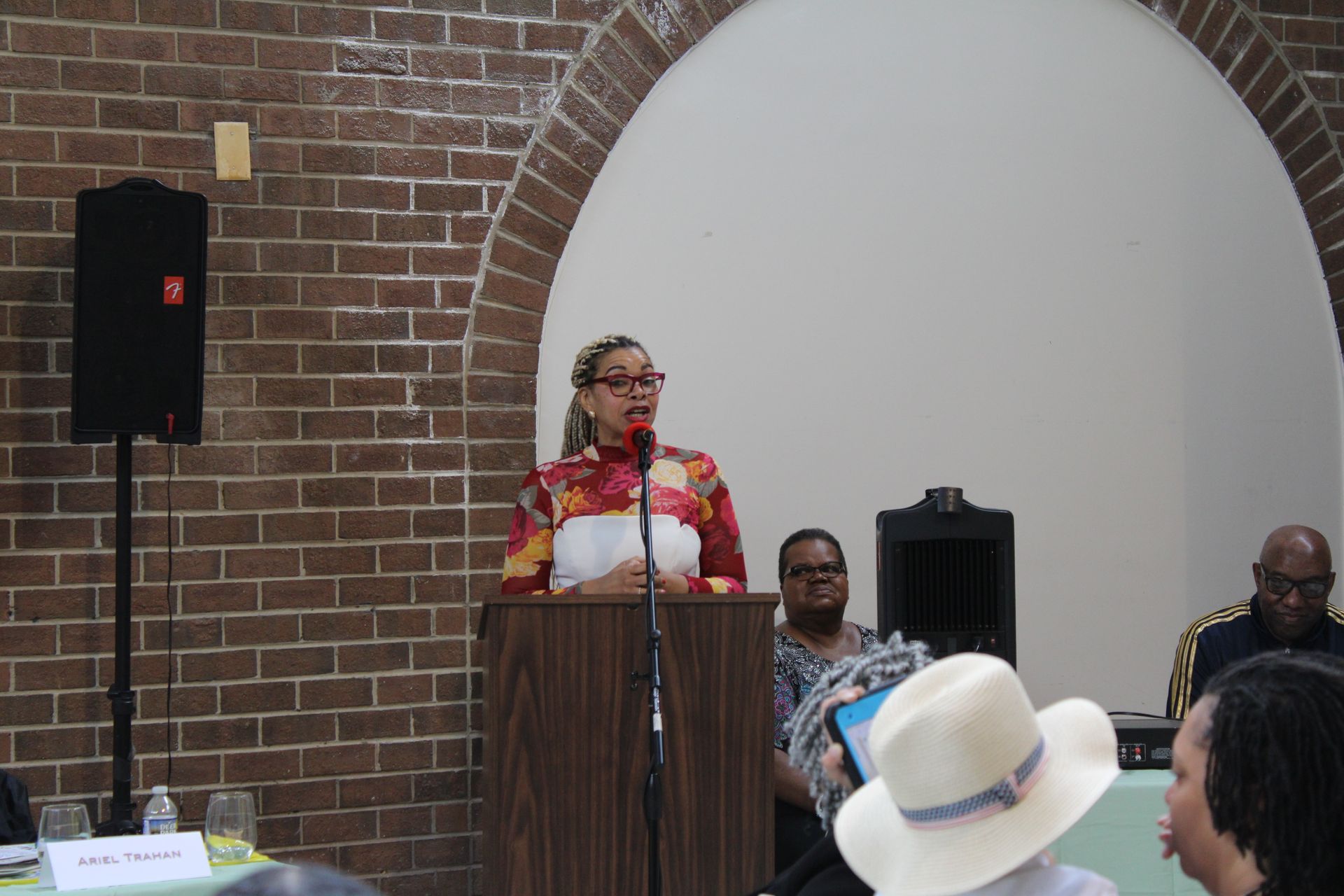 Woman speaking at a podium in a room with an arched doorway. Audience members are visible.