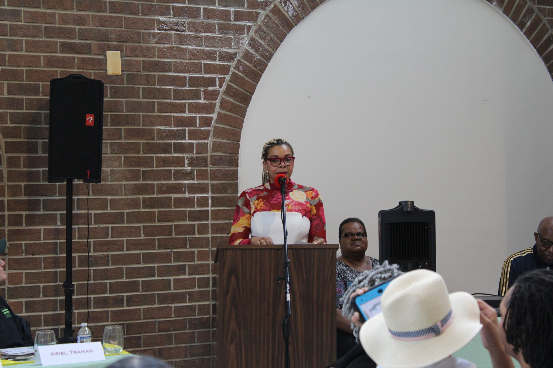 Woman speaking at a podium in a brick-walled room. Audience members seated, speaker with patterned shirt.