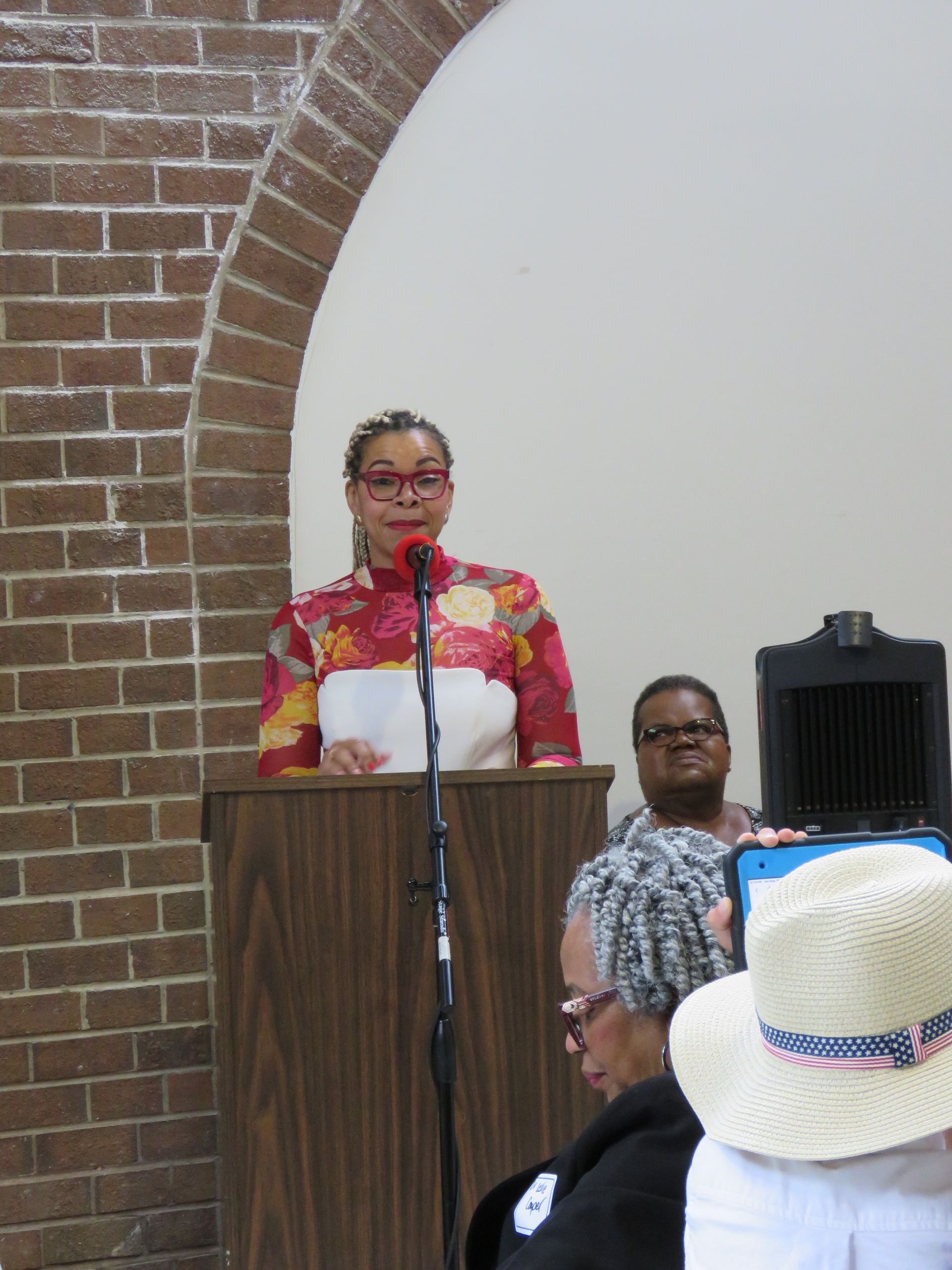 Woman speaking at a podium with a floral top, bricks on the left, and audience in front.