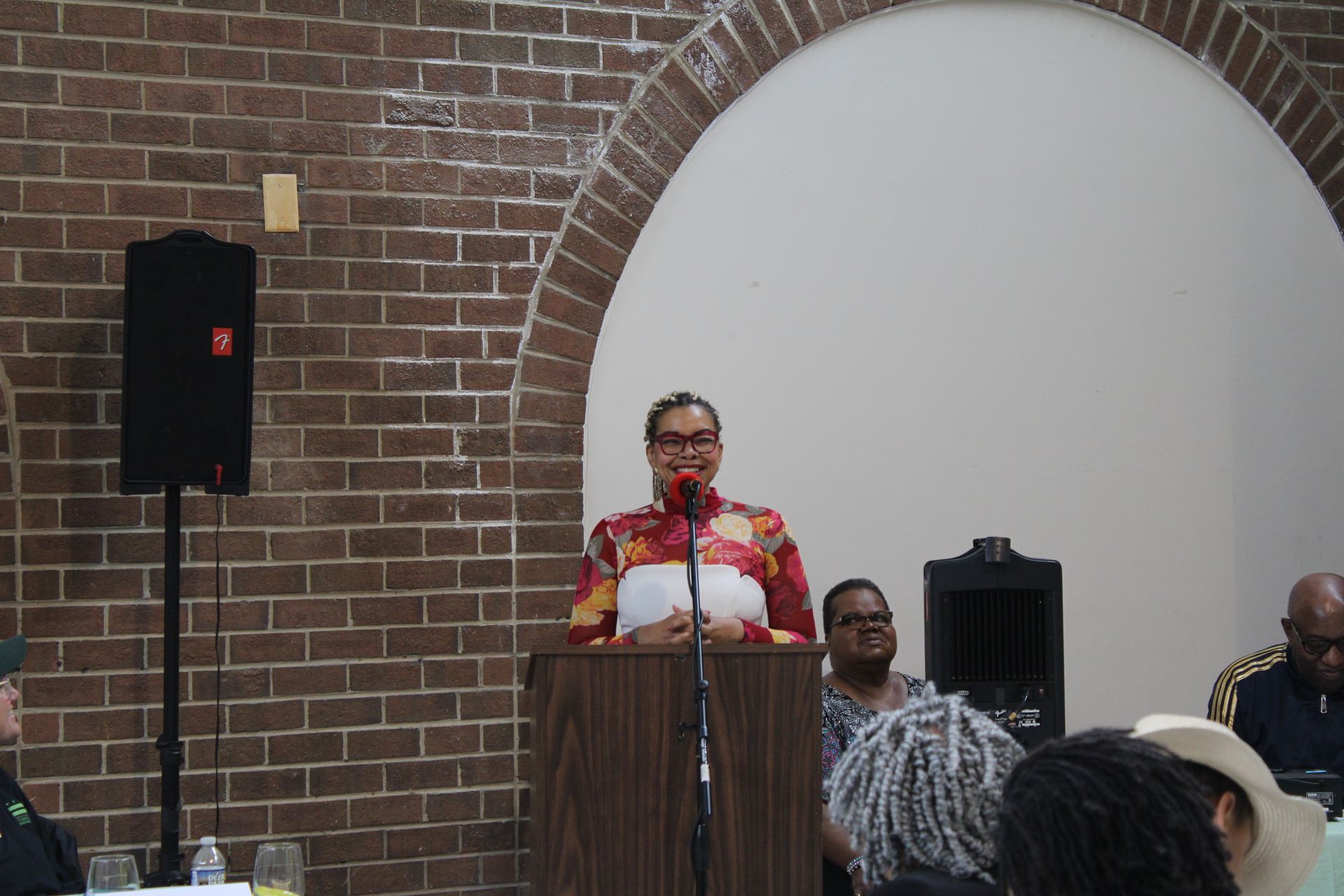 Woman speaking from a podium at an event, brick wall backdrop, archway.