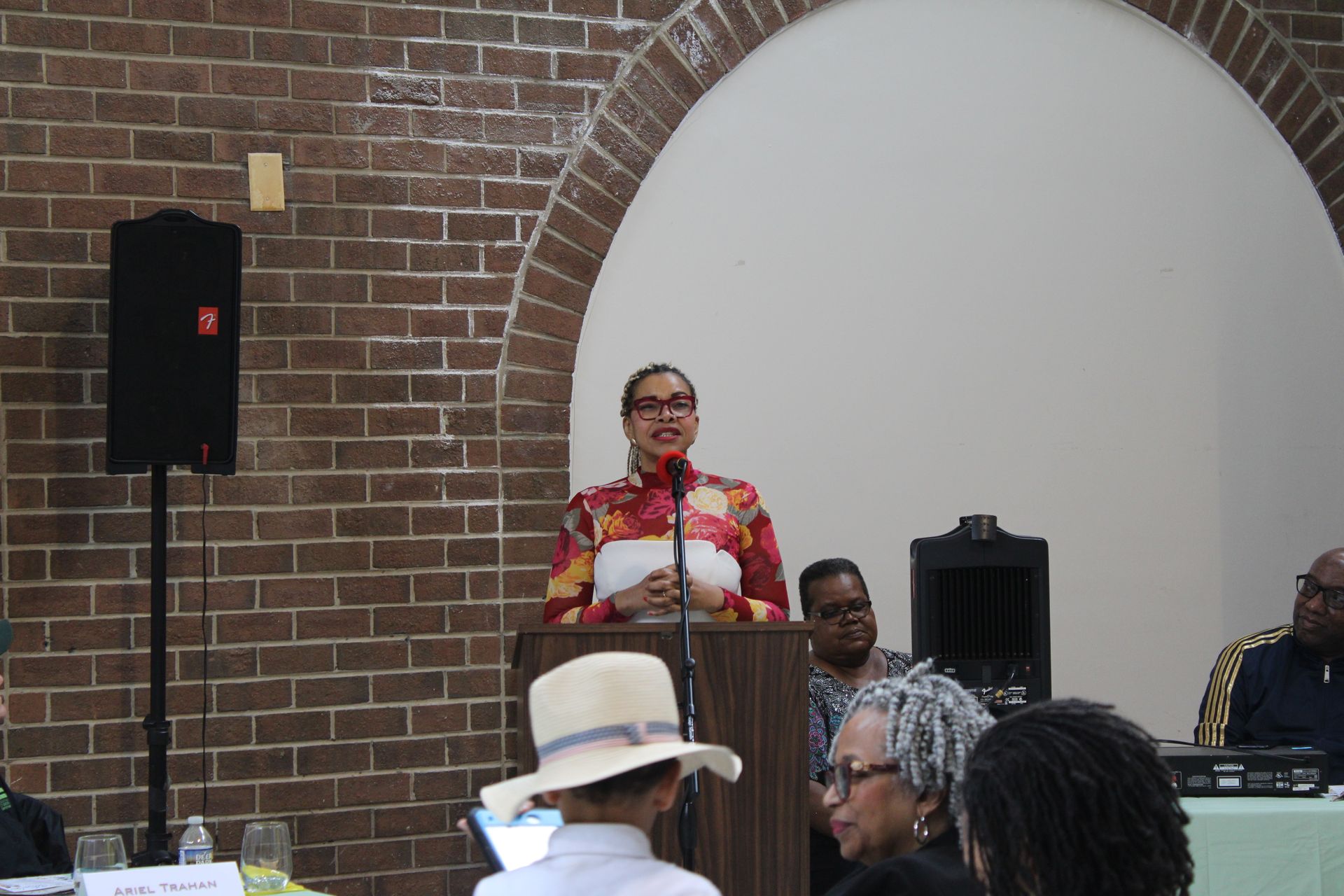 Woman speaking at a podium in a brick-walled room. Audience members seated, listening.