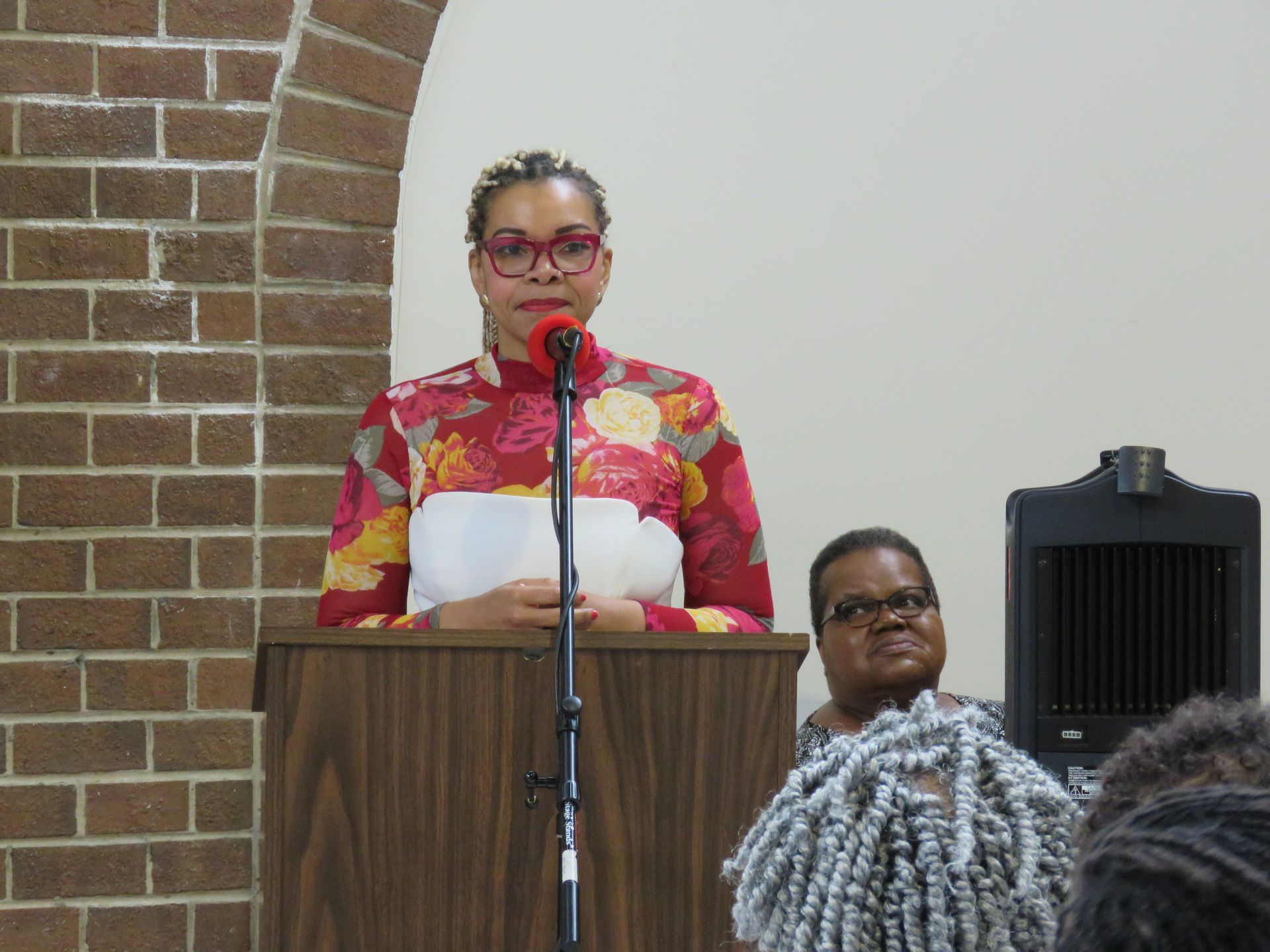 Woman speaking at a podium with a floral top, another woman looks on, brick wall in background.
