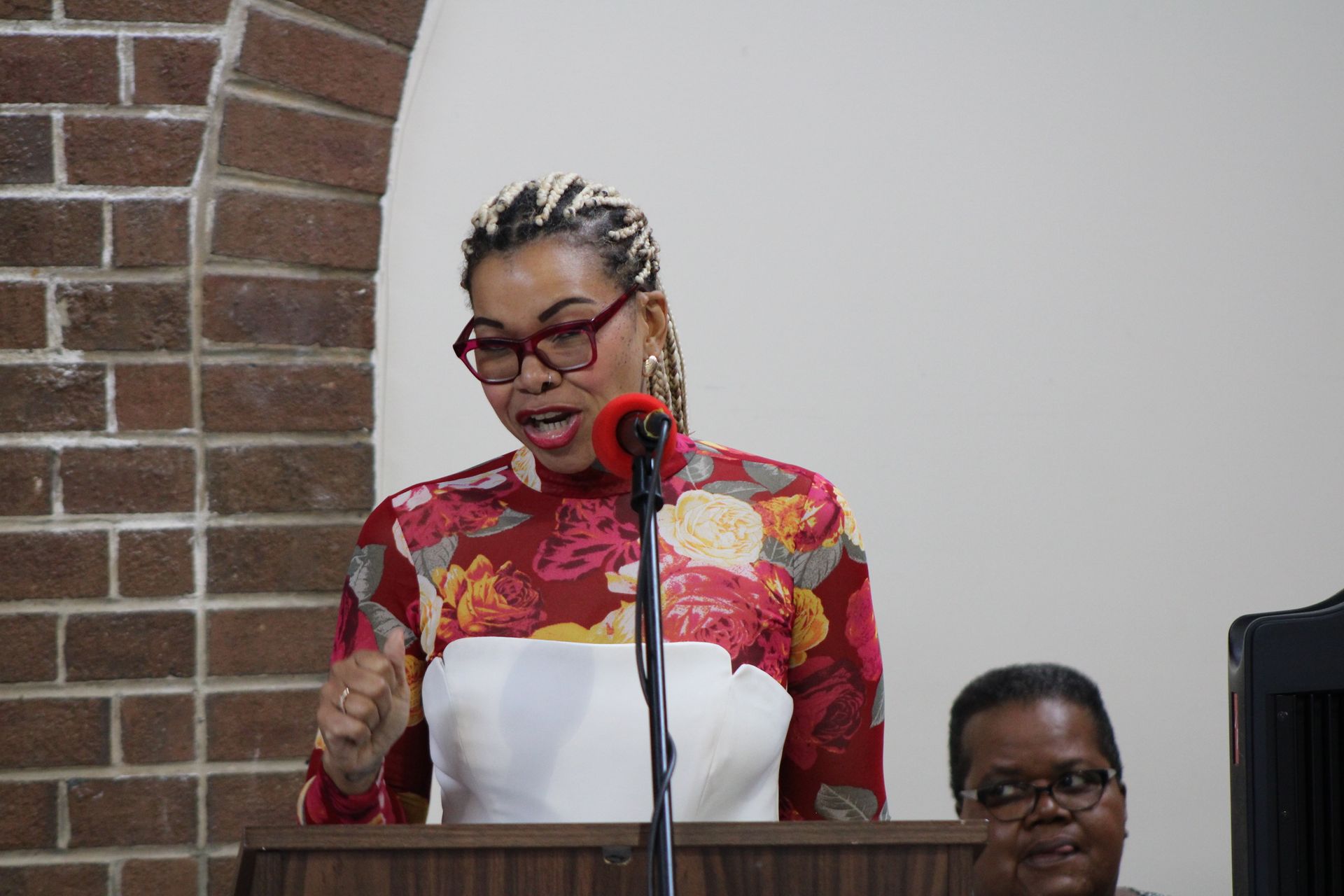 Woman speaking at a lectern with a microphone in front of a brick and white background.