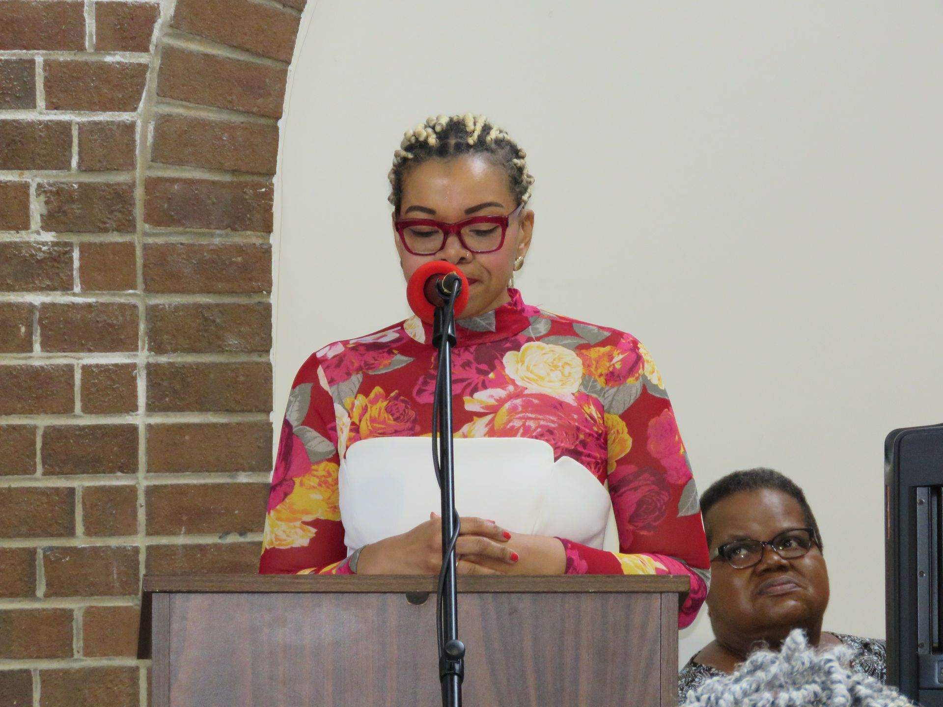 Woman speaking at a lectern with microphone, floral shirt, red glasses, in a brick and white setting.