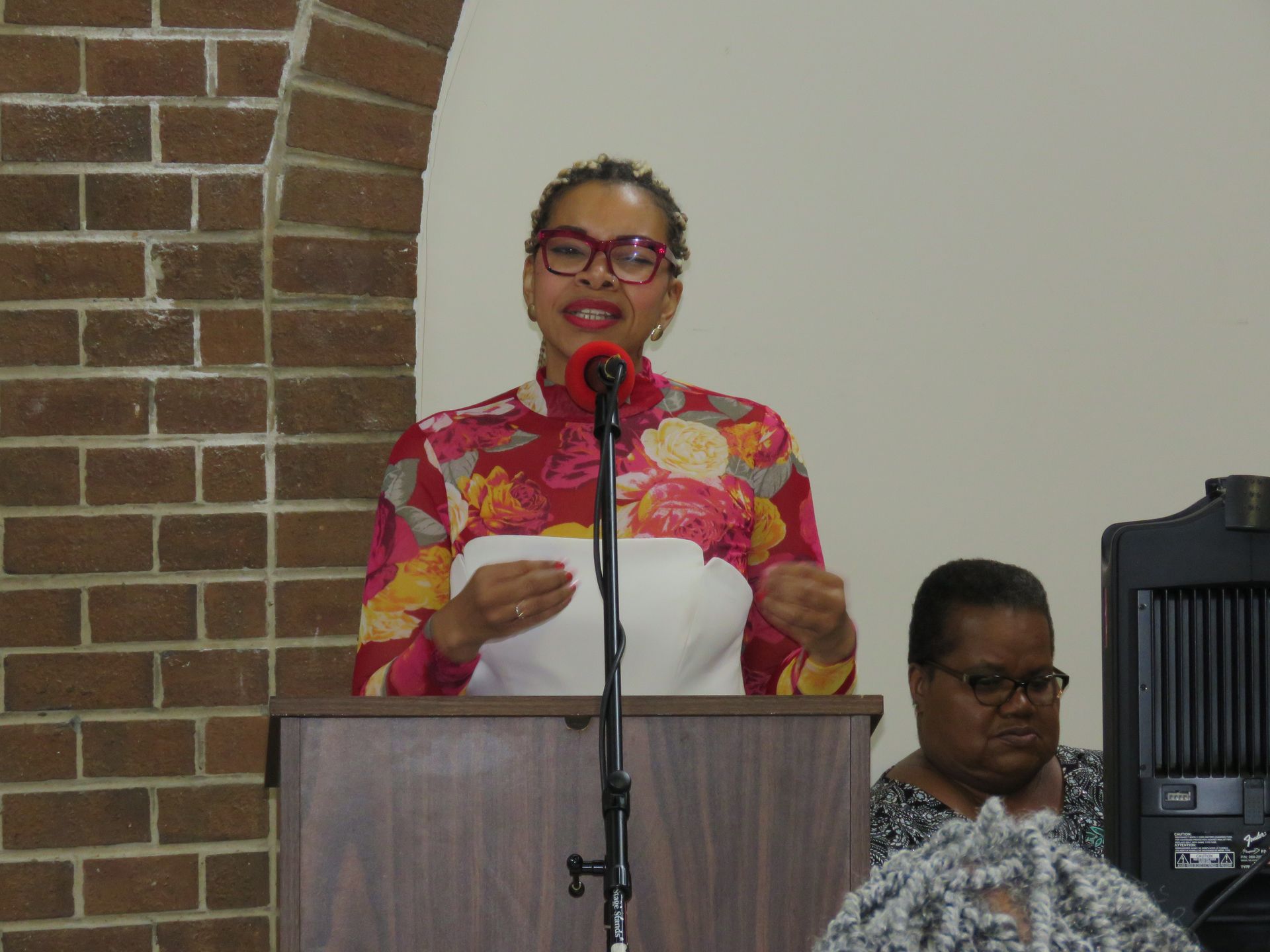 Woman speaking at a podium, floral top, brick wall background, another woman seated beside speaker.