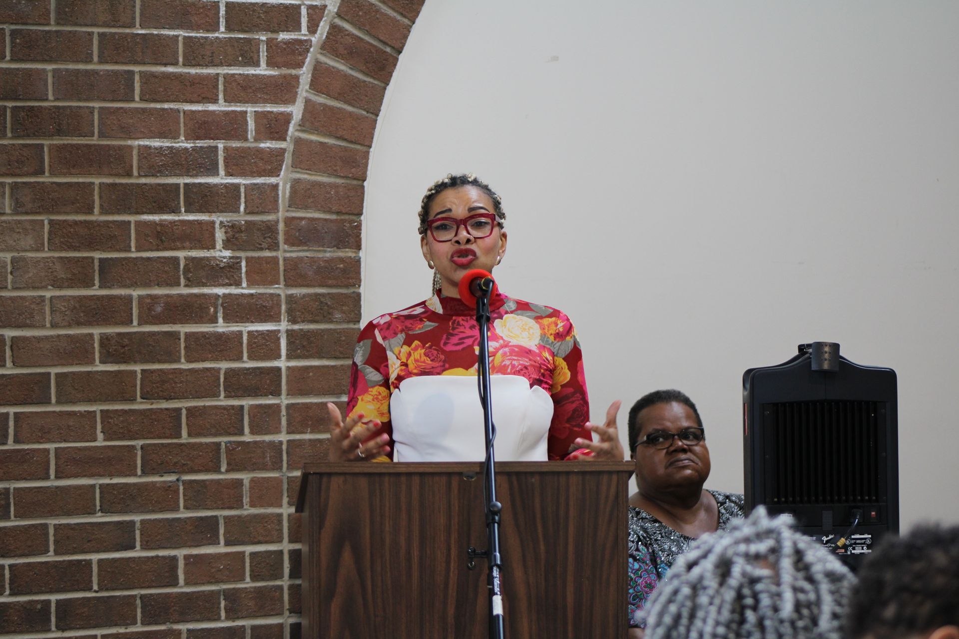 Woman speaking at a podium in a brick-walled room; another woman sits behind her, and audience members are visible.