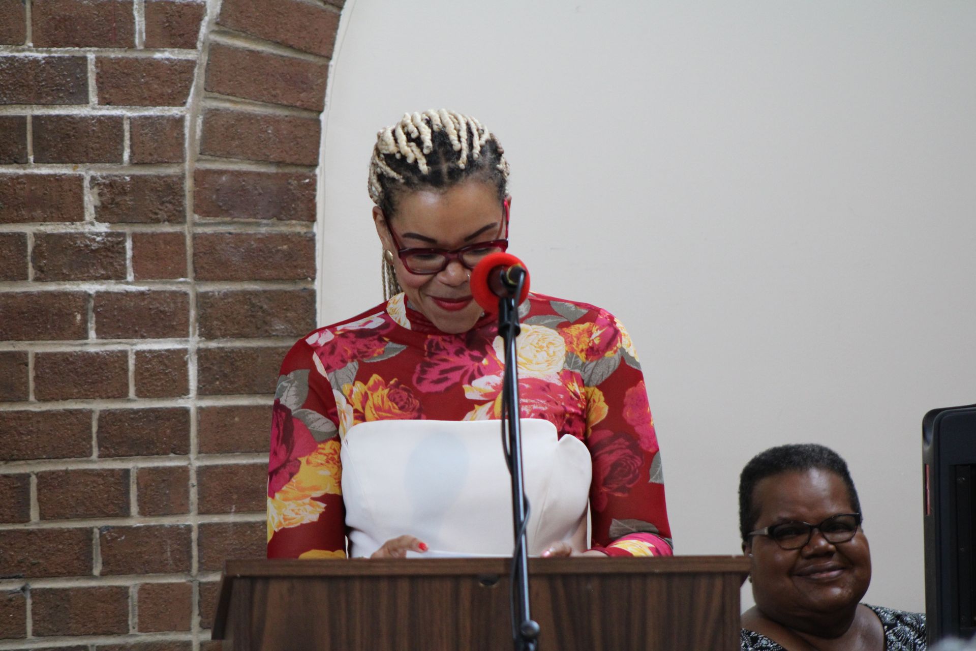 Woman speaking at a lectern with microphone, reading from papers, brick wall in background.