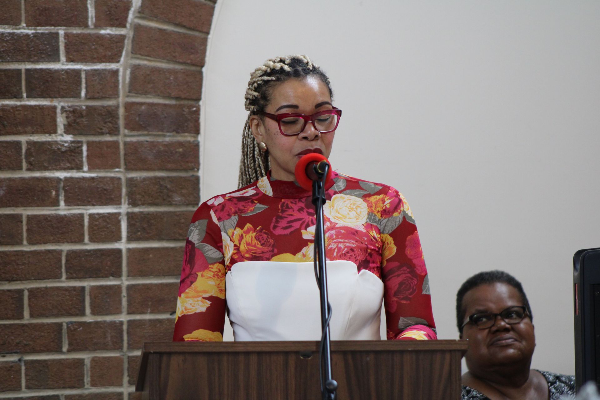 Woman speaking at a podium, wearing glasses and floral top, with brick wall background.