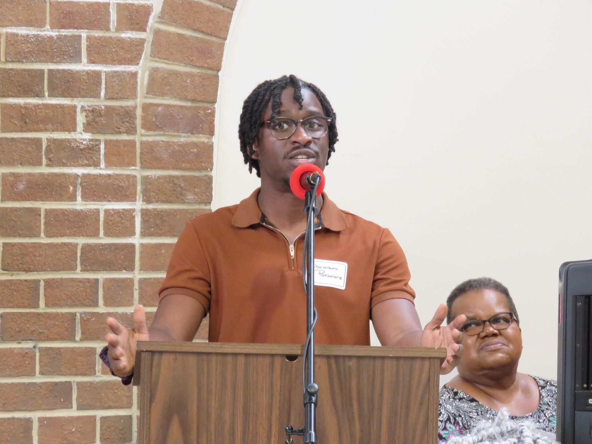 Man speaking at a podium with arms raised. Brick wall and another person are in the background.