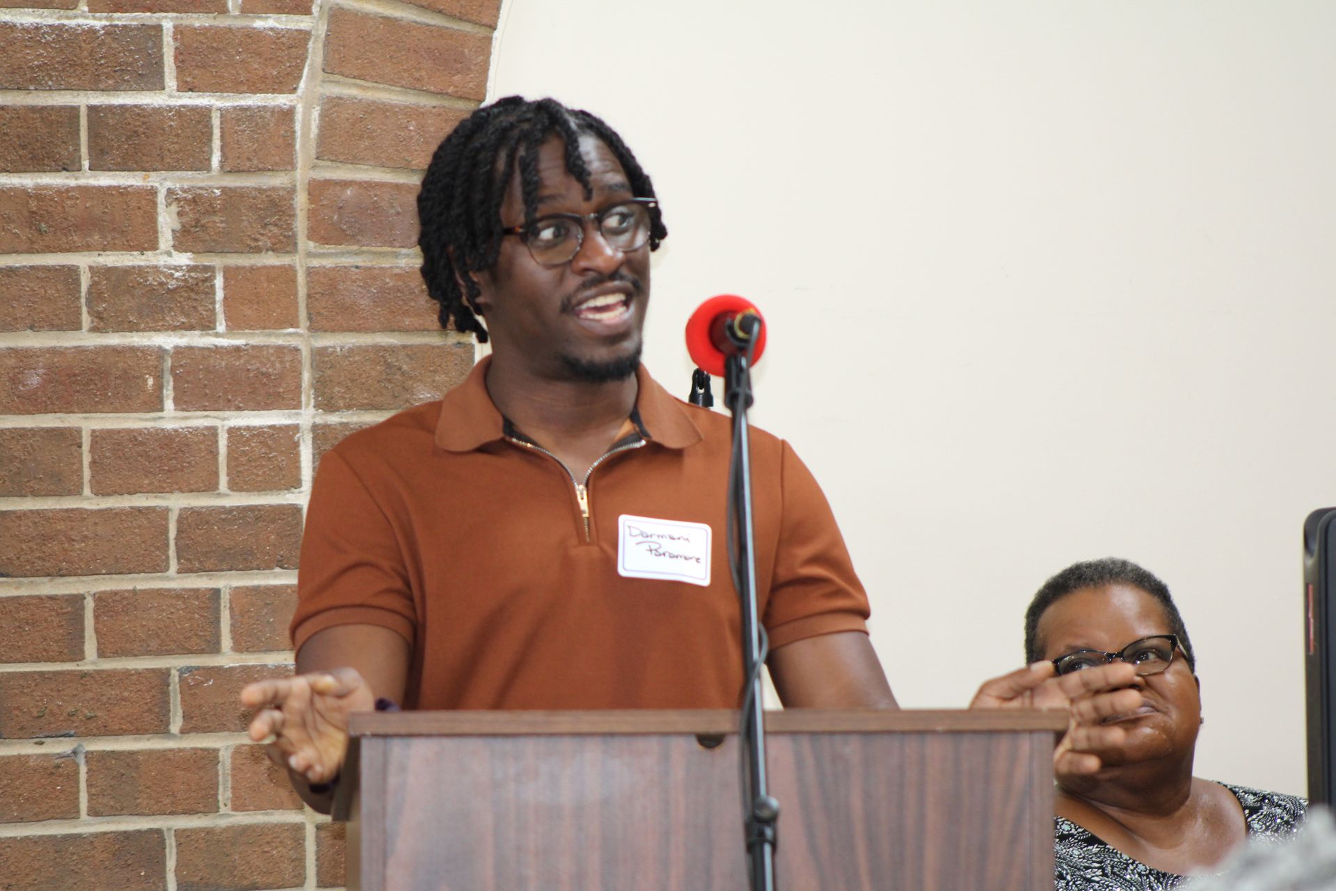 Man speaking at a podium with a brick wall backdrop. Woman in background.